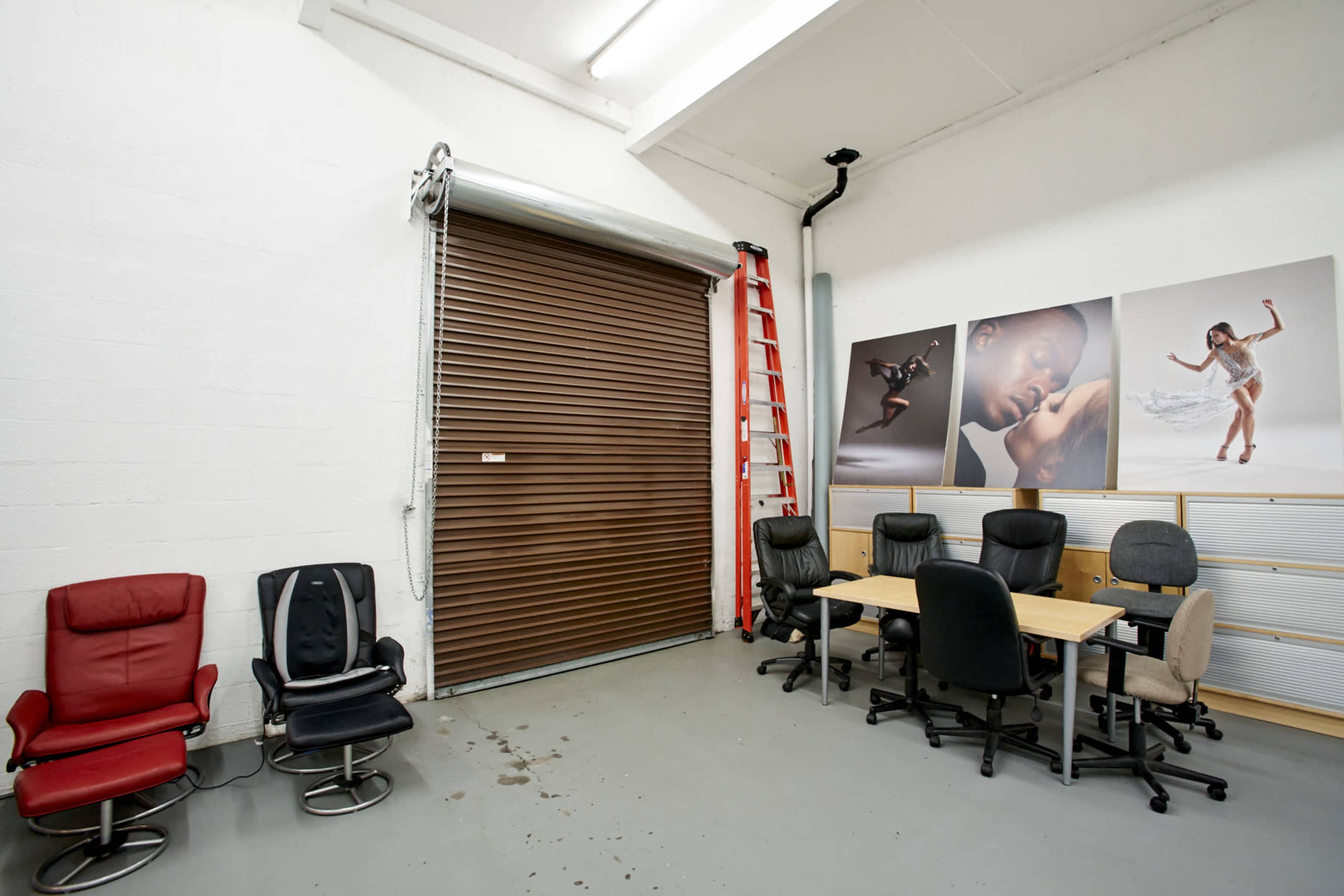 A spacious room with a metal roll-up door, a red ladder, a set of office chairs, and a table arrangement against the wall, featuring large photographs.