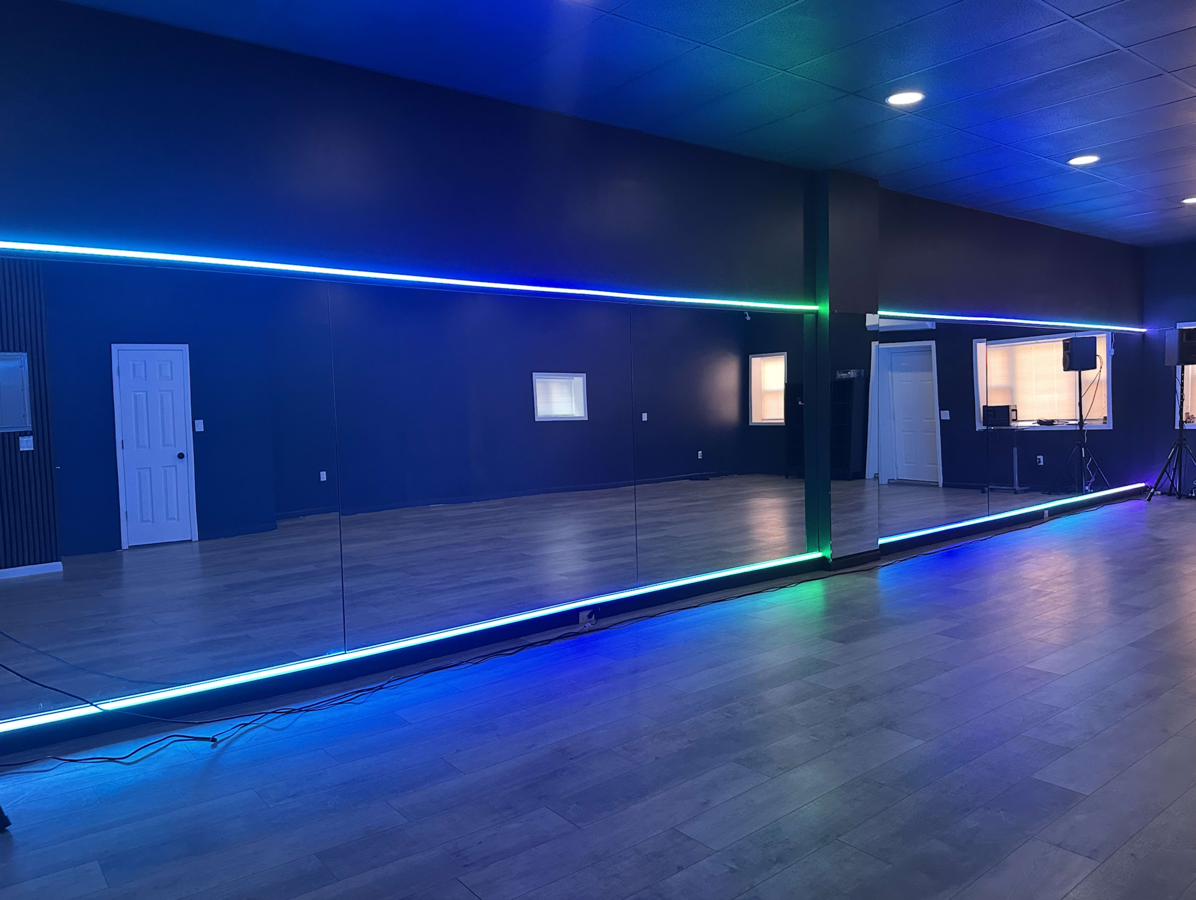A spacious dance studio features a long mirror along one wall, illuminated by colorful LED strip lights.