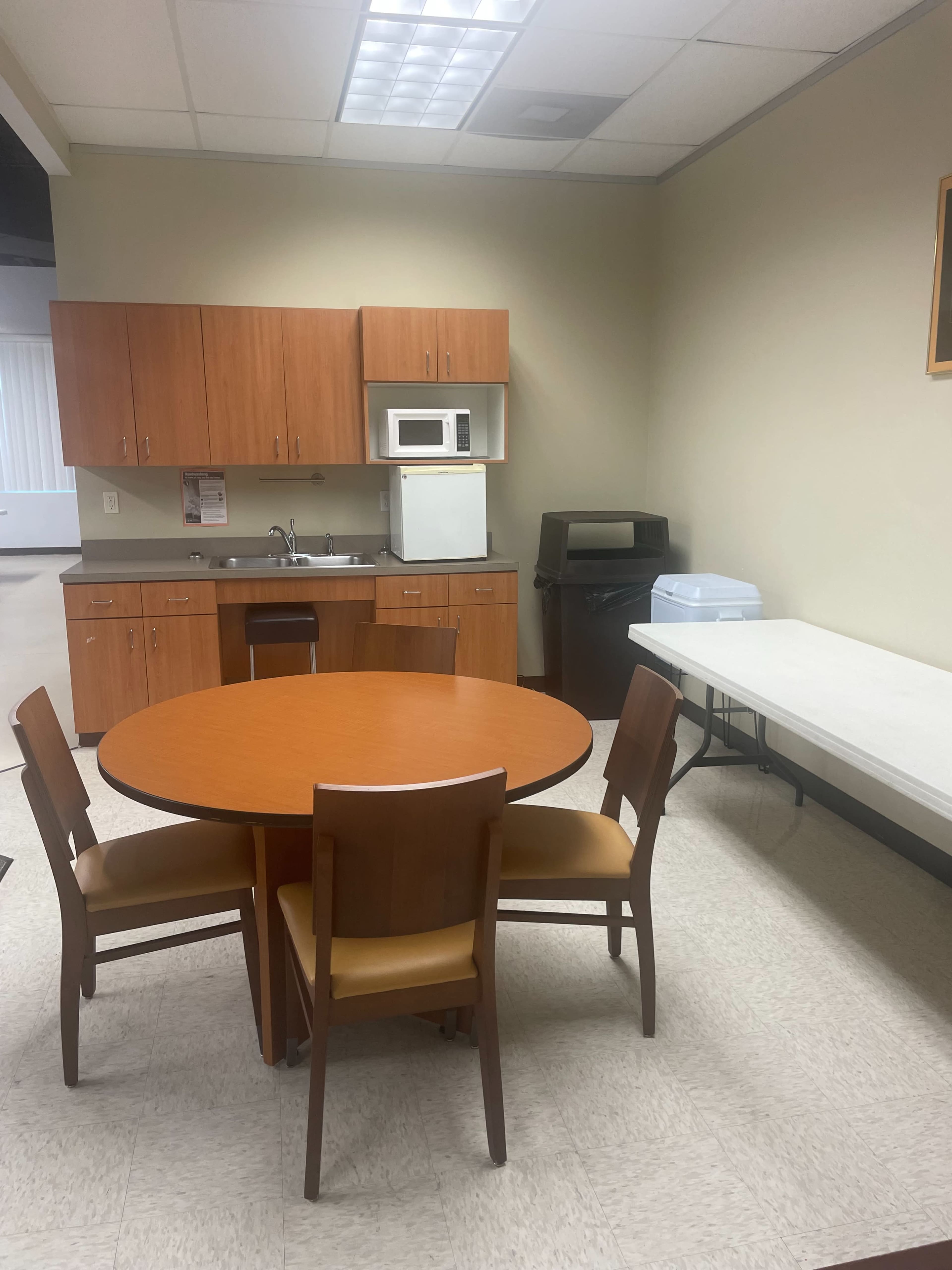 A small break room with a round table and four chairs, a kitchenette with cabinets, a microwave, and a countertop, along with a folding table against the wall.