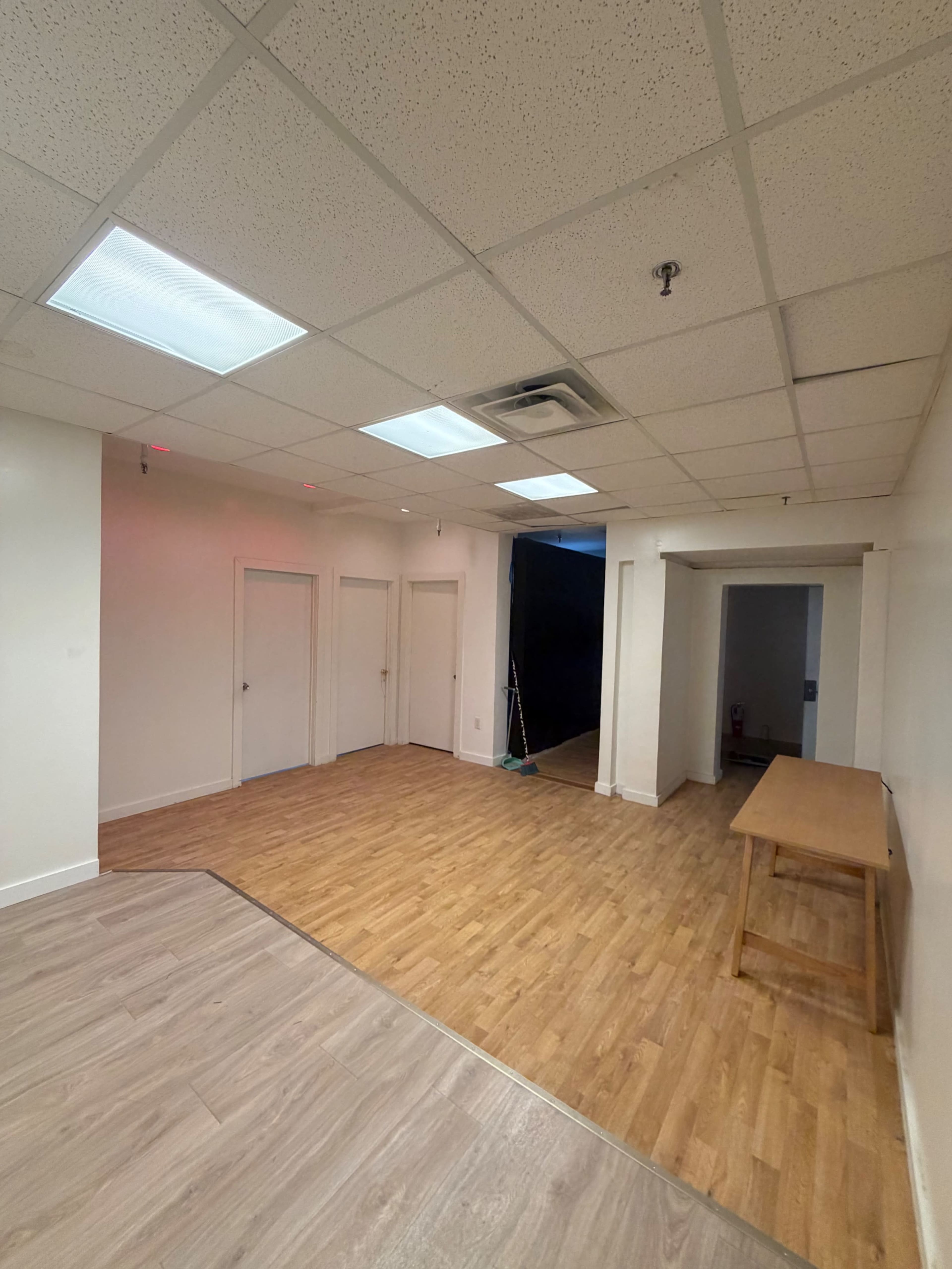 The image shows a minimally furnished interior space with wooden flooring, several closed doors, and a table against one wall.