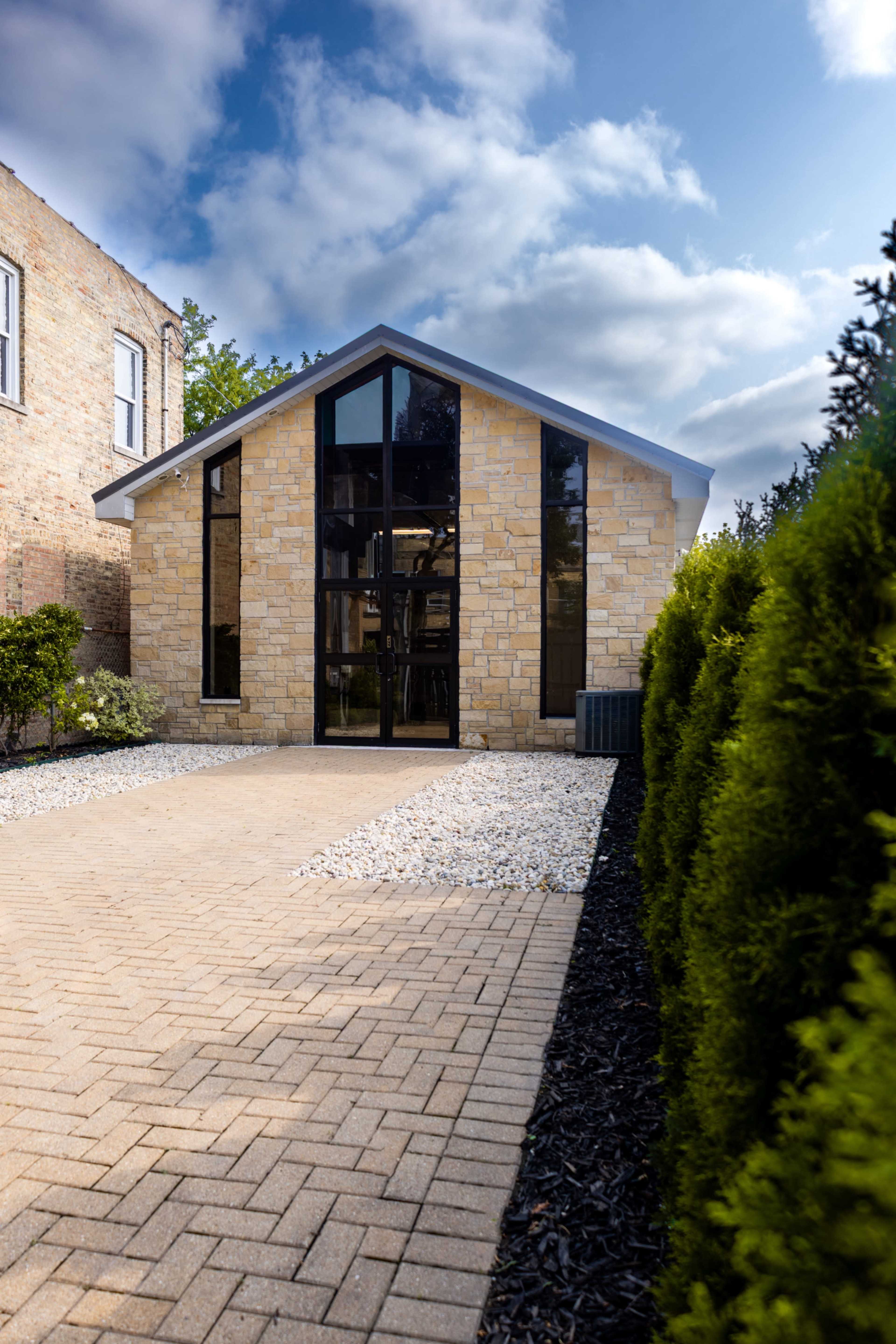 A modern stone house with large windows is set in a landscaped area featuring a paved driveway and greenery on either side.