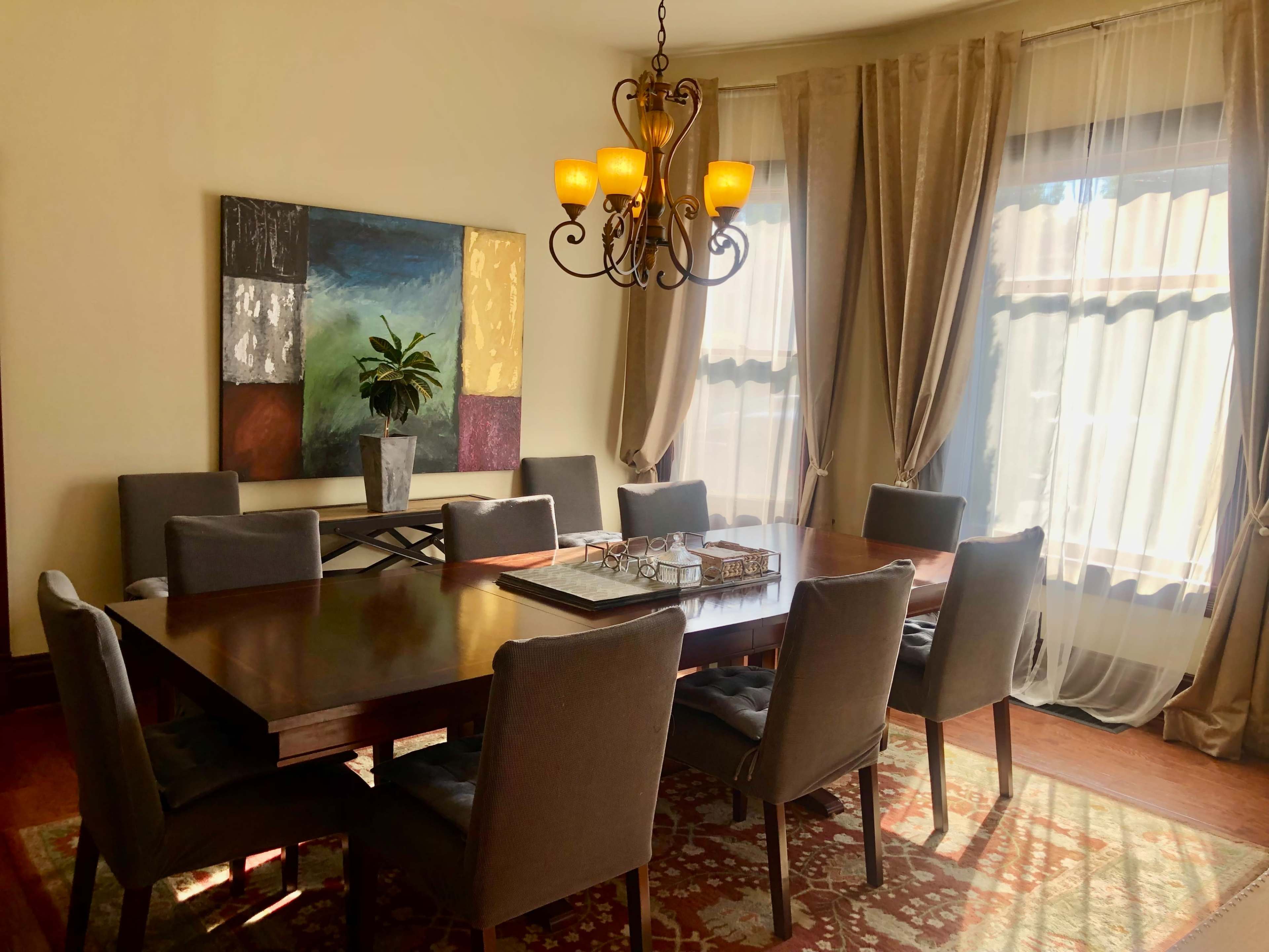 A dining room features a large wooden table surrounded by gray upholstered chairs, with a chandelier above and a colorful abstract painting on the wall.