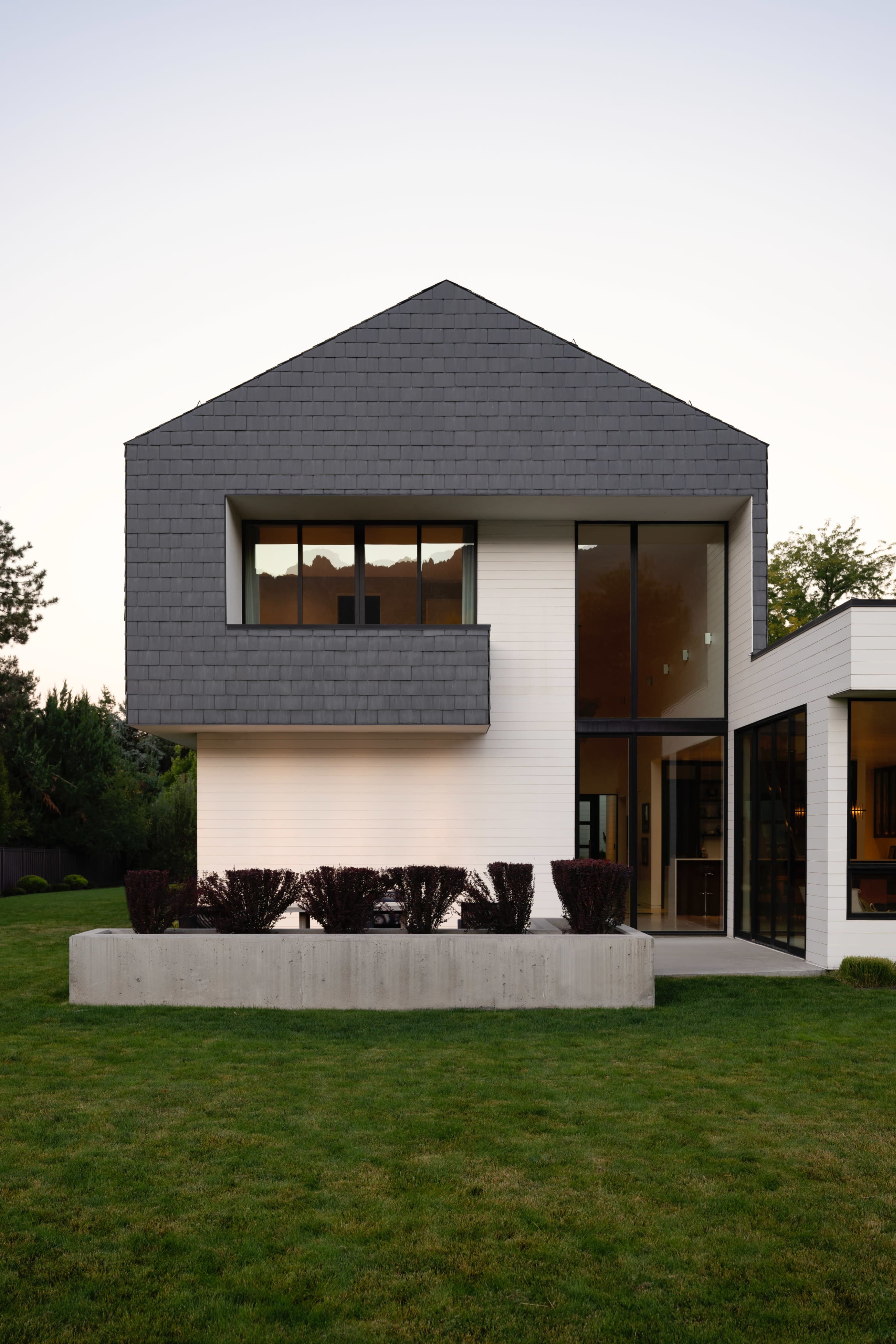 The image shows a modern two-story house with a geometric design, featuring a gray roof, large windows, and a landscaped yard with low shrubs.