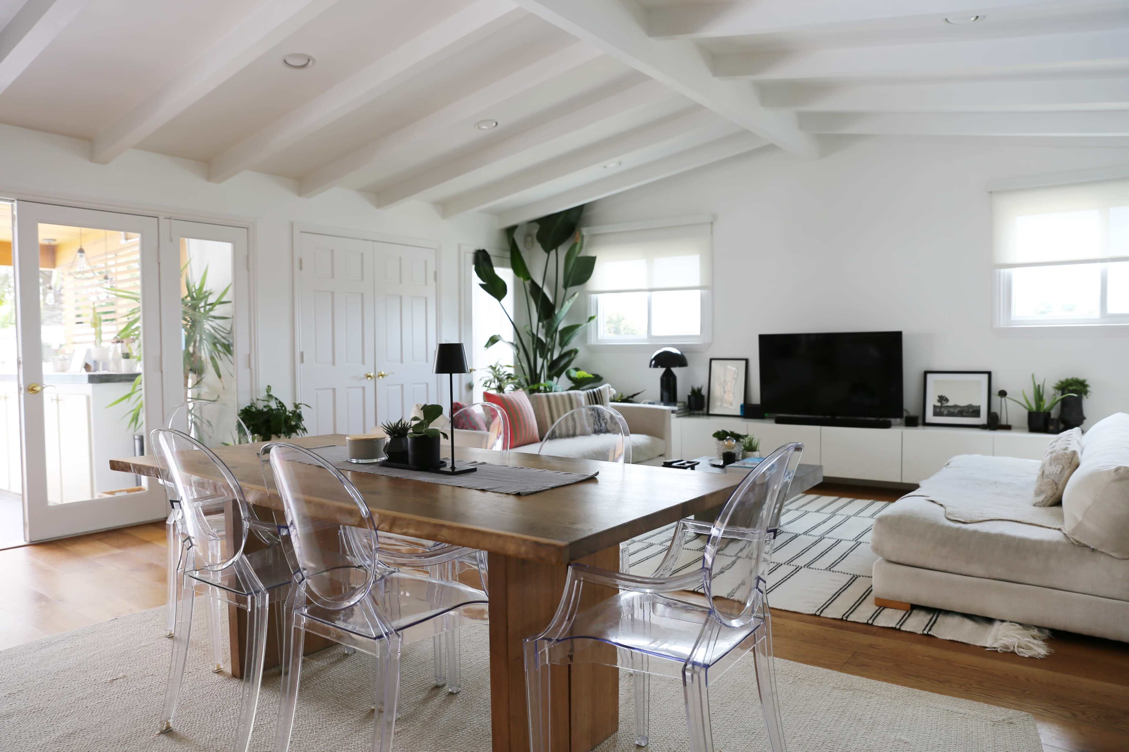 A bright, modern living and dining area featuring a wooden table, clear acrylic chairs, a large television, and various indoor plants.