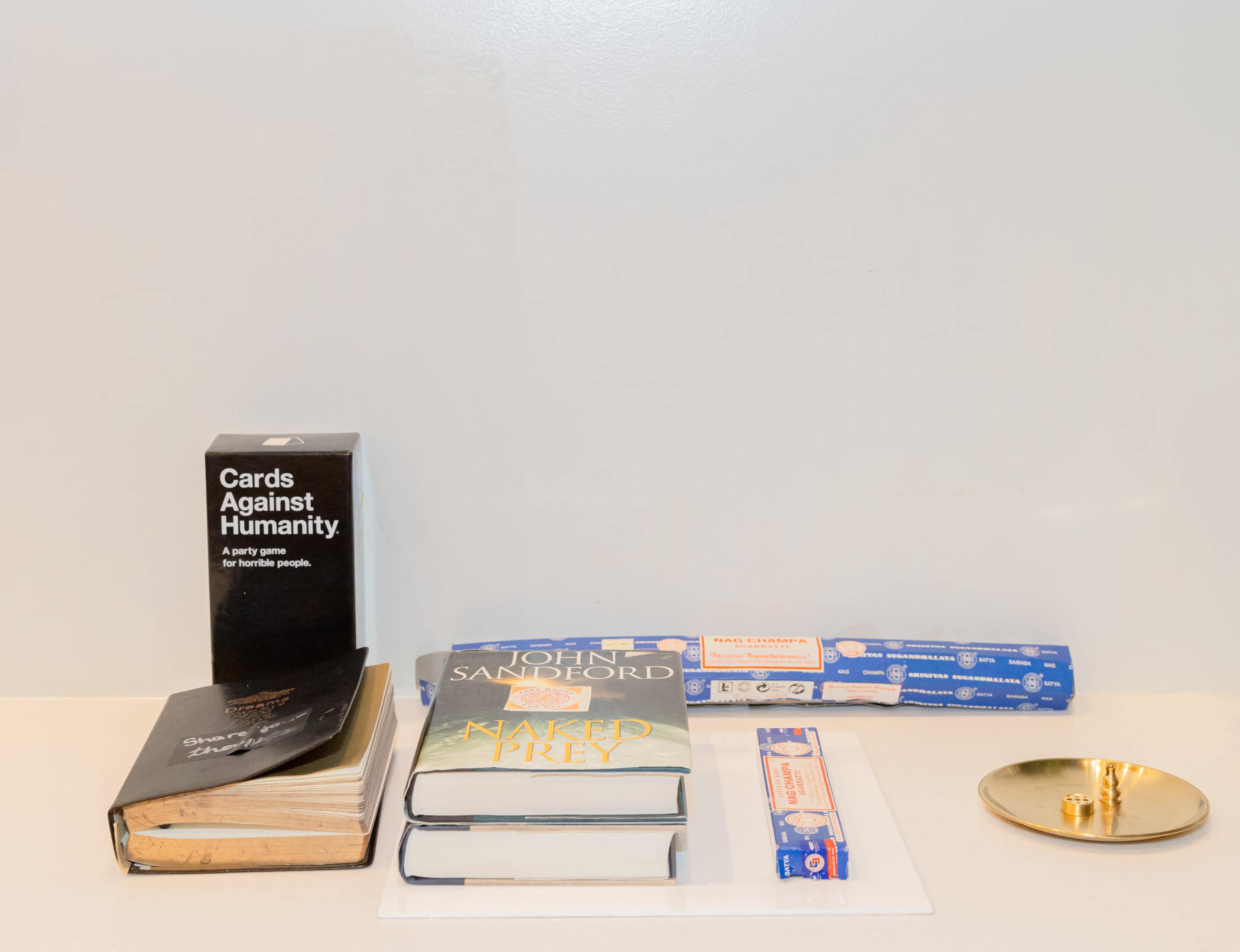 A tabletop arrangement that includes a box of "Cards Against Humanity," three stacked books, a roll of pasta, and a gold-colored dish.