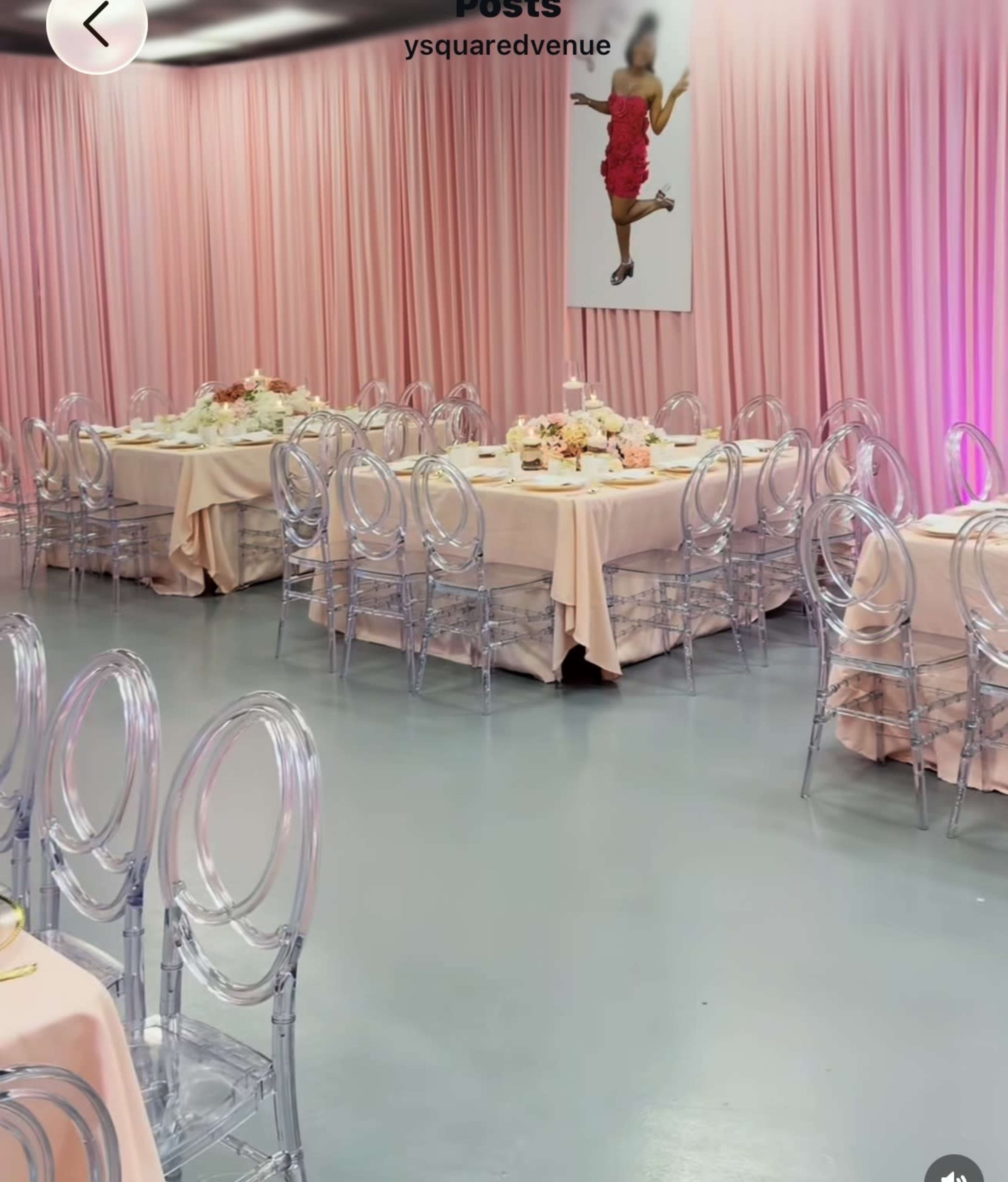 The image shows a stylish event space with pink draped walls, clear acrylic chairs, and tables set with floral centerpieces.