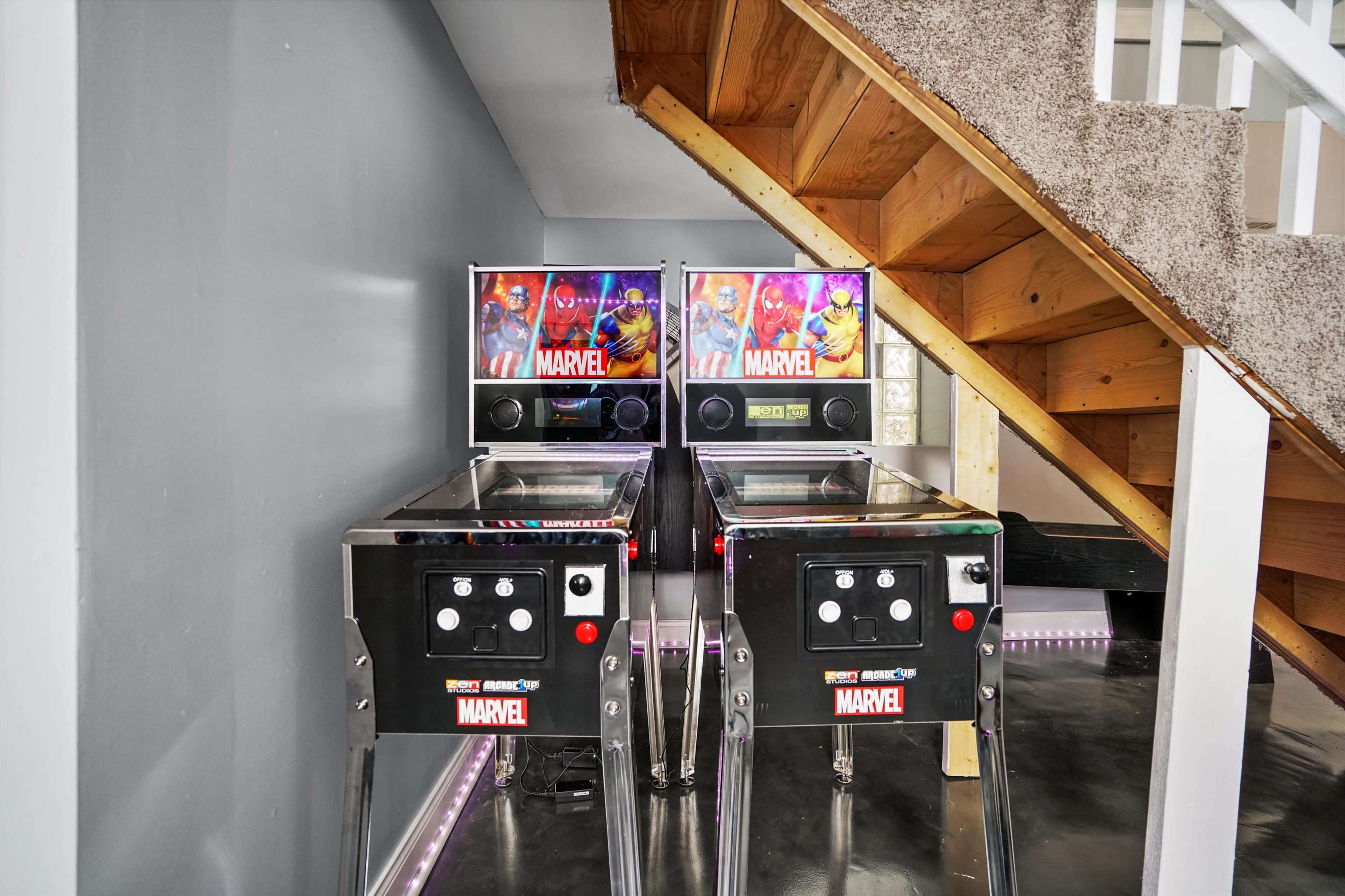 Two Marvel-themed pinball machines are positioned against a gray wall beneath a staircase.