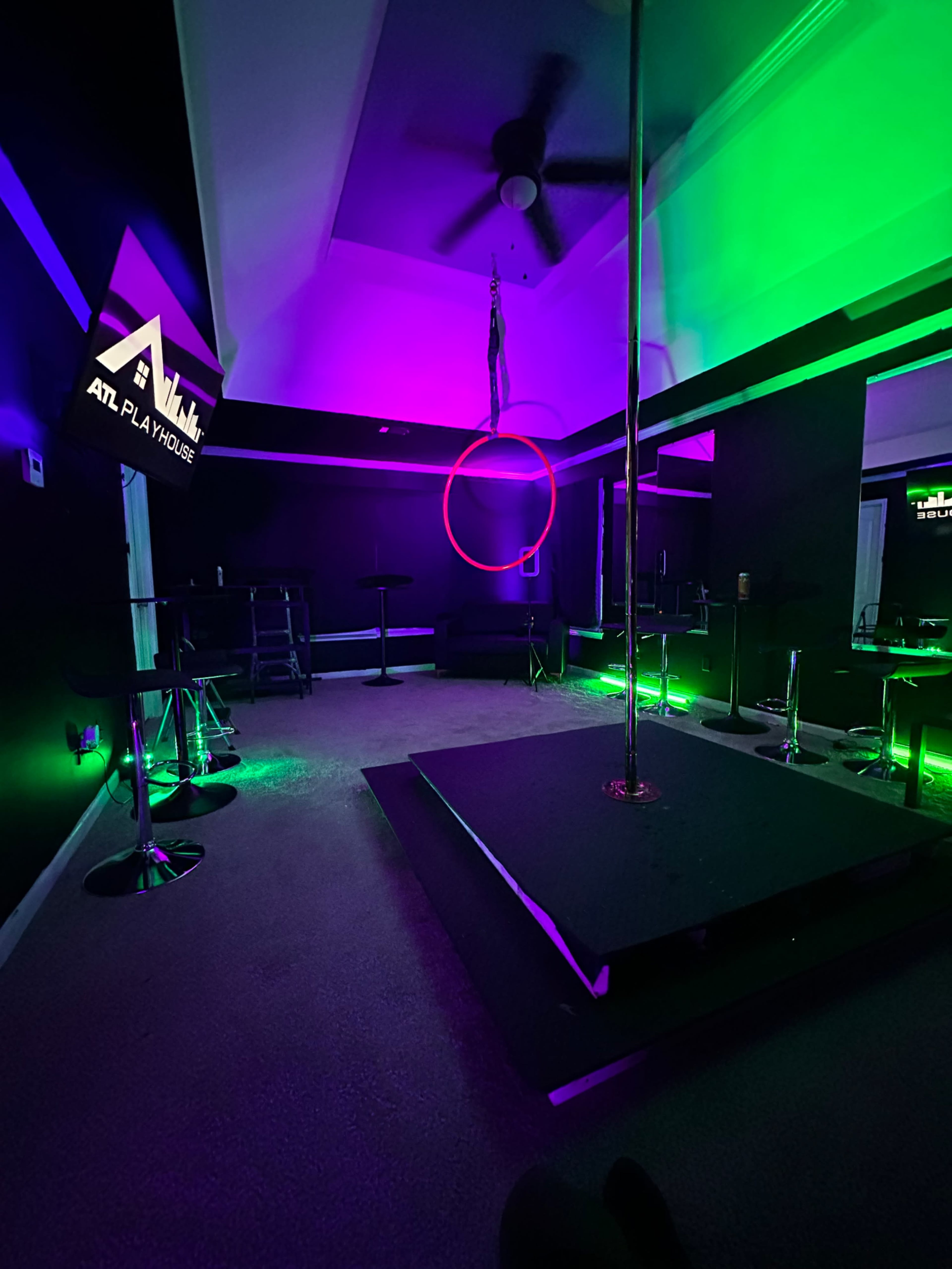 The image shows a dimly lit room with a central pole, colorful neon lights in green and purple, bar stools, and a sign indicating "ATL Playhouse."