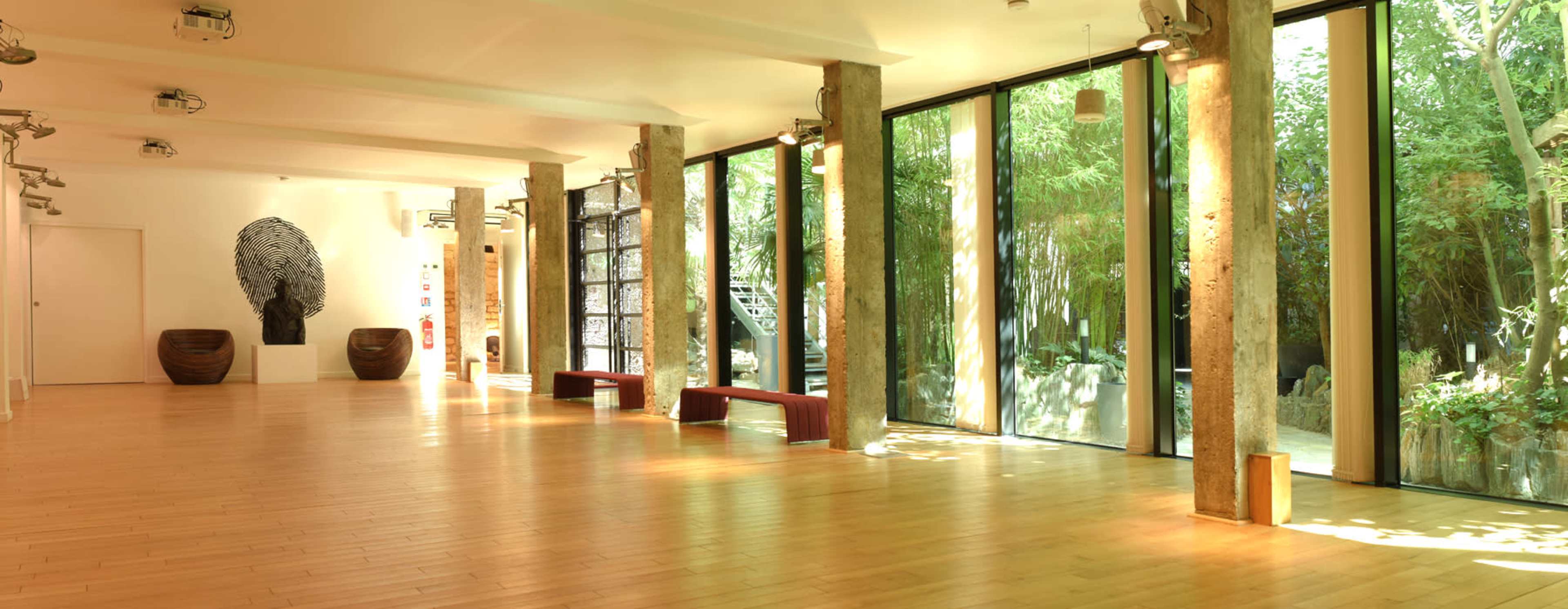 A spacious room with wooden flooring, large glass windows, and concrete pillars, featuring minimalist decor and greenery visible outside.