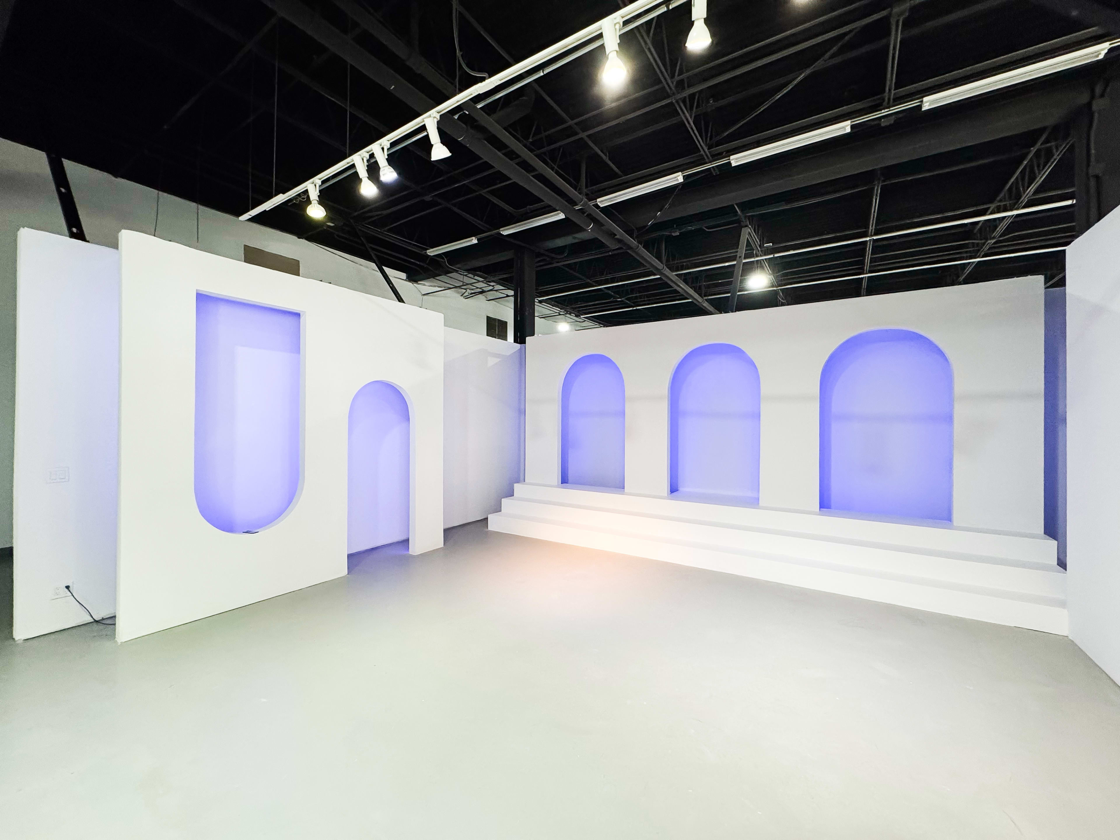 The image shows a minimalist interior space featuring white walls, a stepped platform, and three arched openings illuminated with soft blue light.