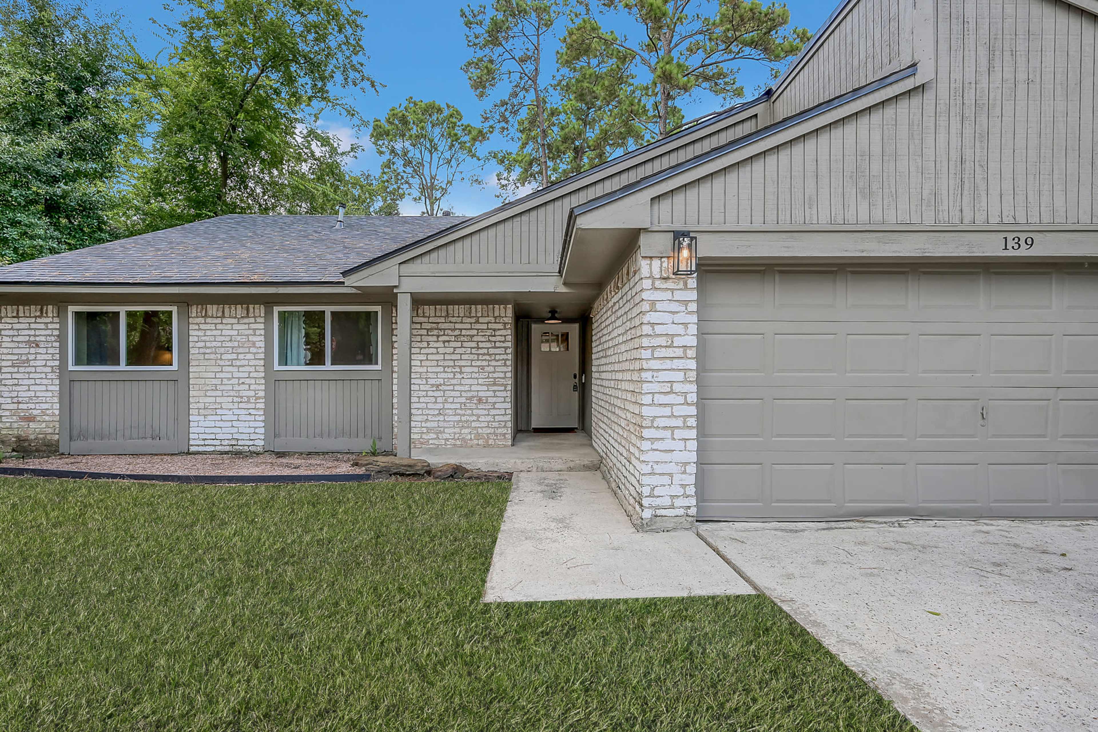 The image shows a single-story house with a light gray exterior, a garage door, and a front entrance framed by trees and landscaped grass.