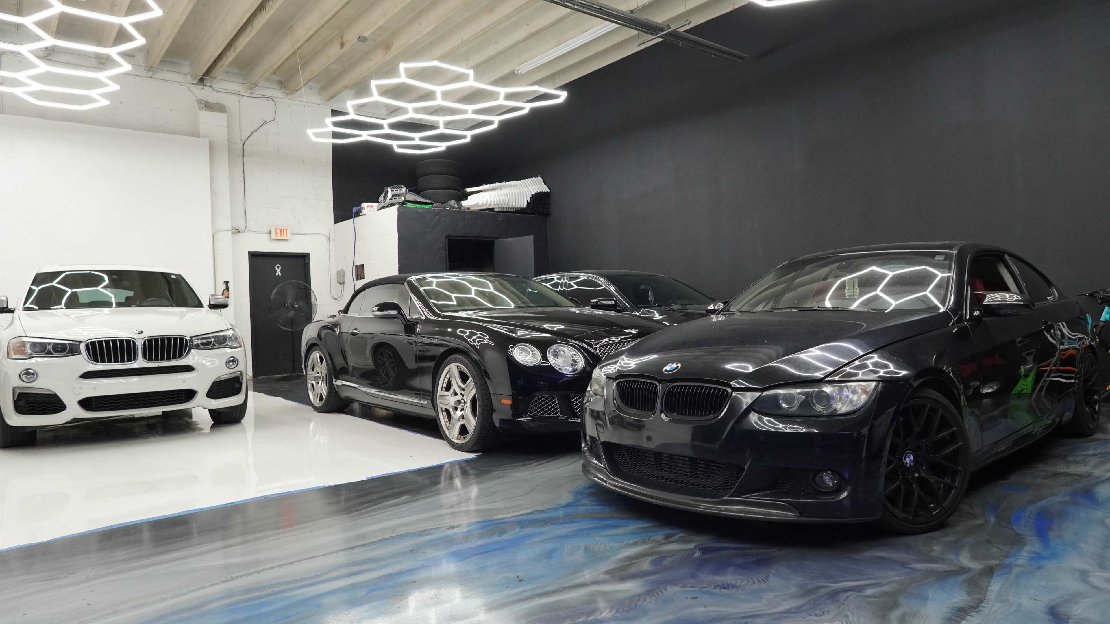 The image shows a modern garage with three luxury cars: a white SUV, a black coupe, and a black sedan, parked on a glossy floor beneath geometric lighting.