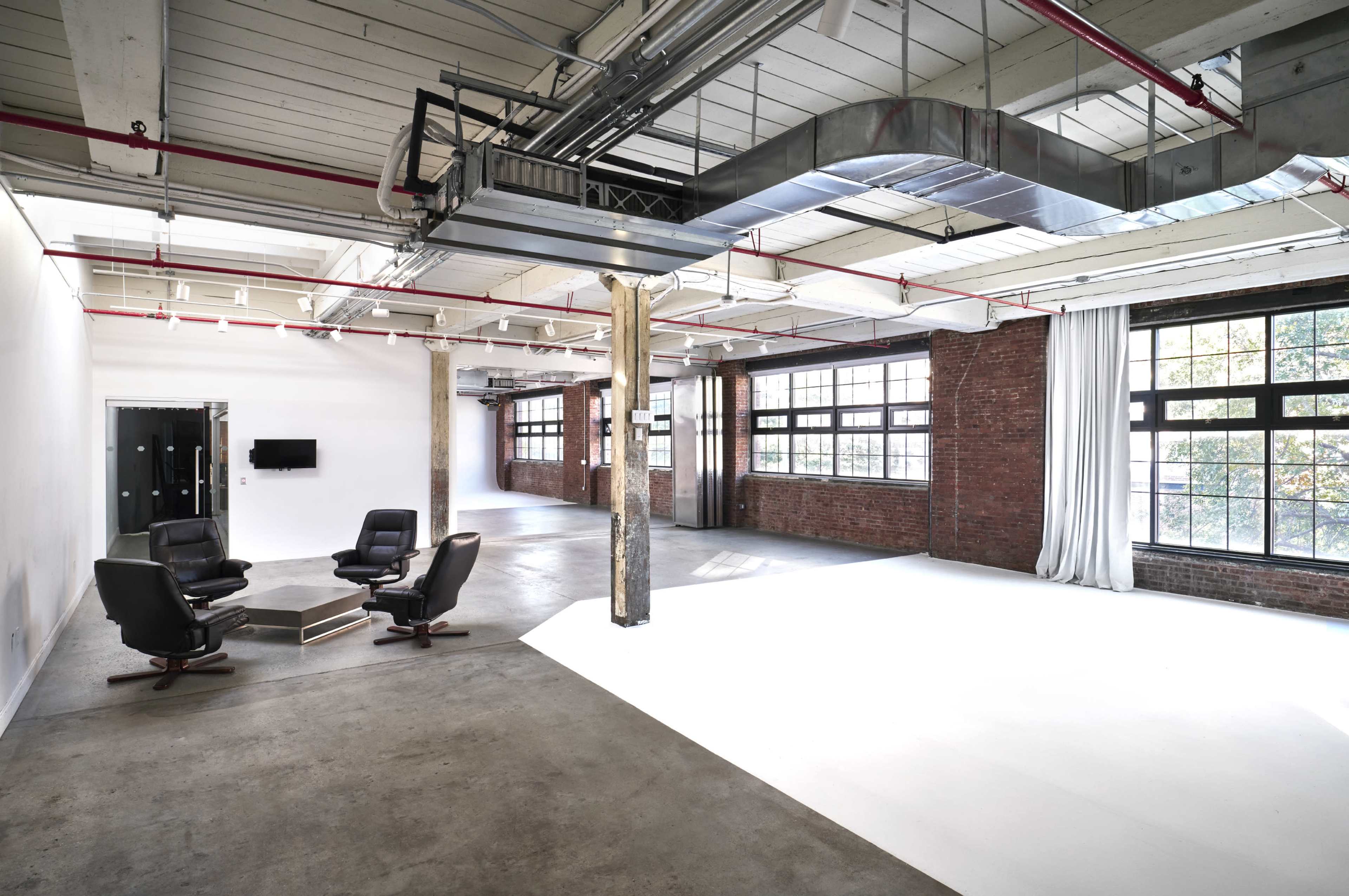 An industrial-style loft with exposed beams, large windows, and a seating area consisting of four chairs around a central table.