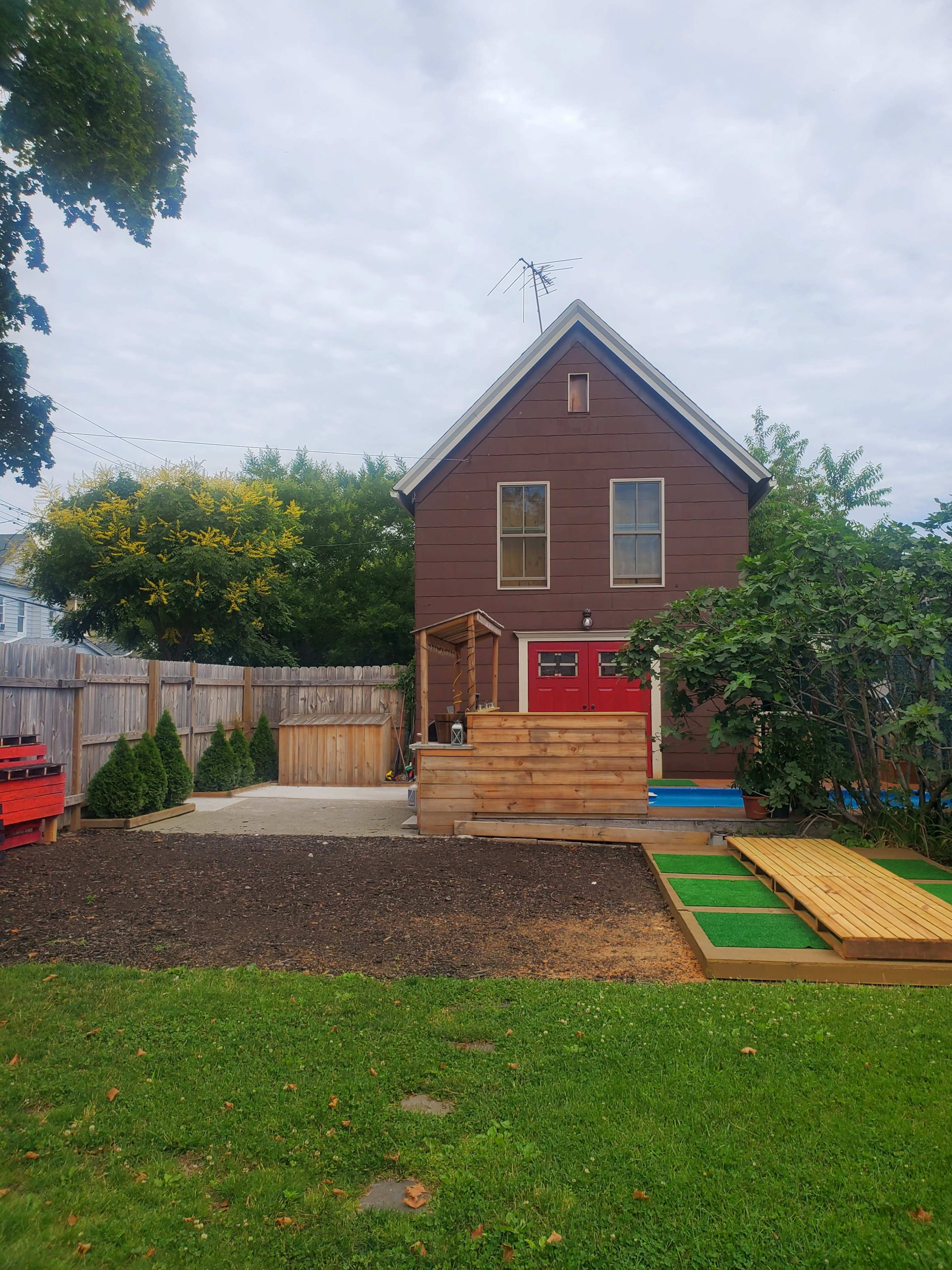 A brown two-story house with a red front door is surrounded by a fenced yard featuring landscaping and a small patio area.