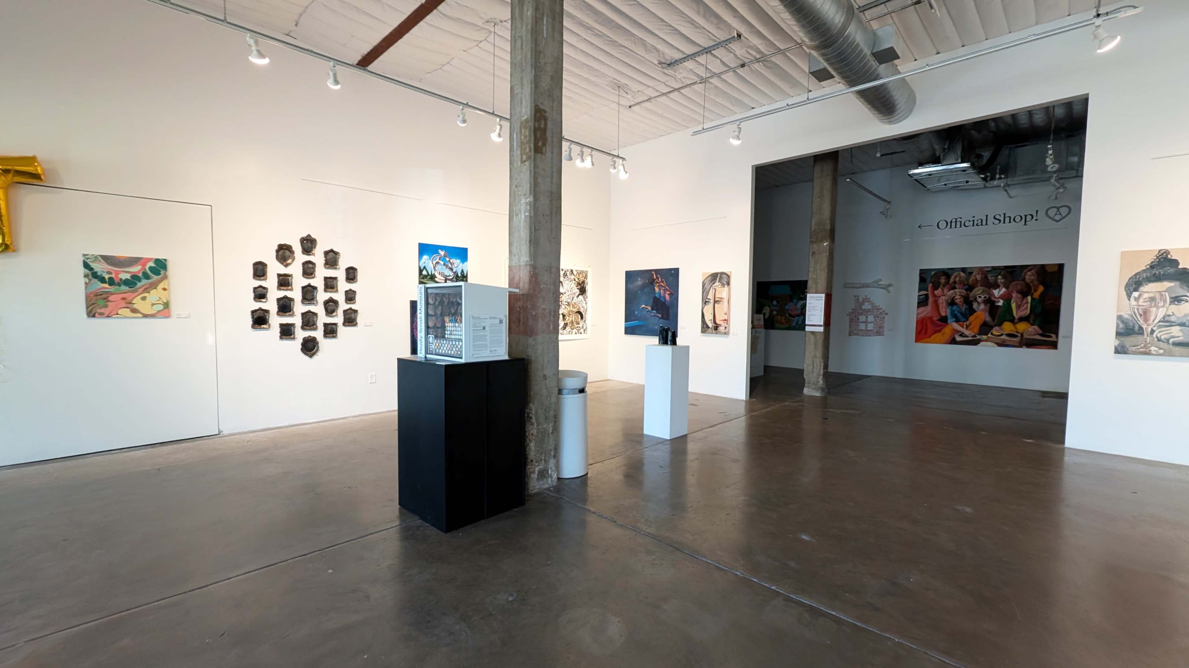 The image shows an art gallery interior featuring various artworks displayed on the walls and on pedestals, with exposed beams and concrete flooring.