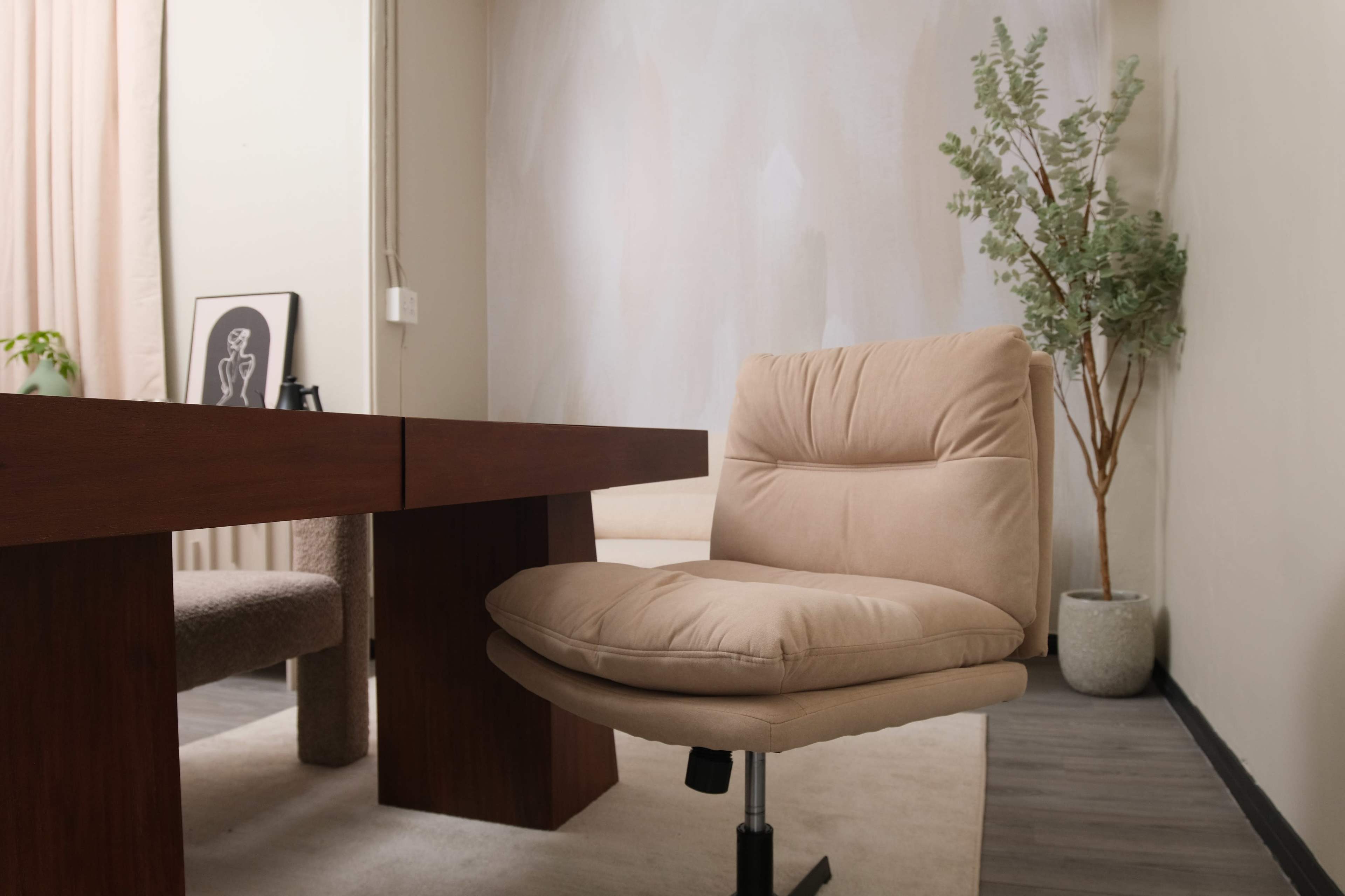 The image shows a minimalist office setting featuring a wooden desk, a beige rolling chair, and a potted plant in the corner.