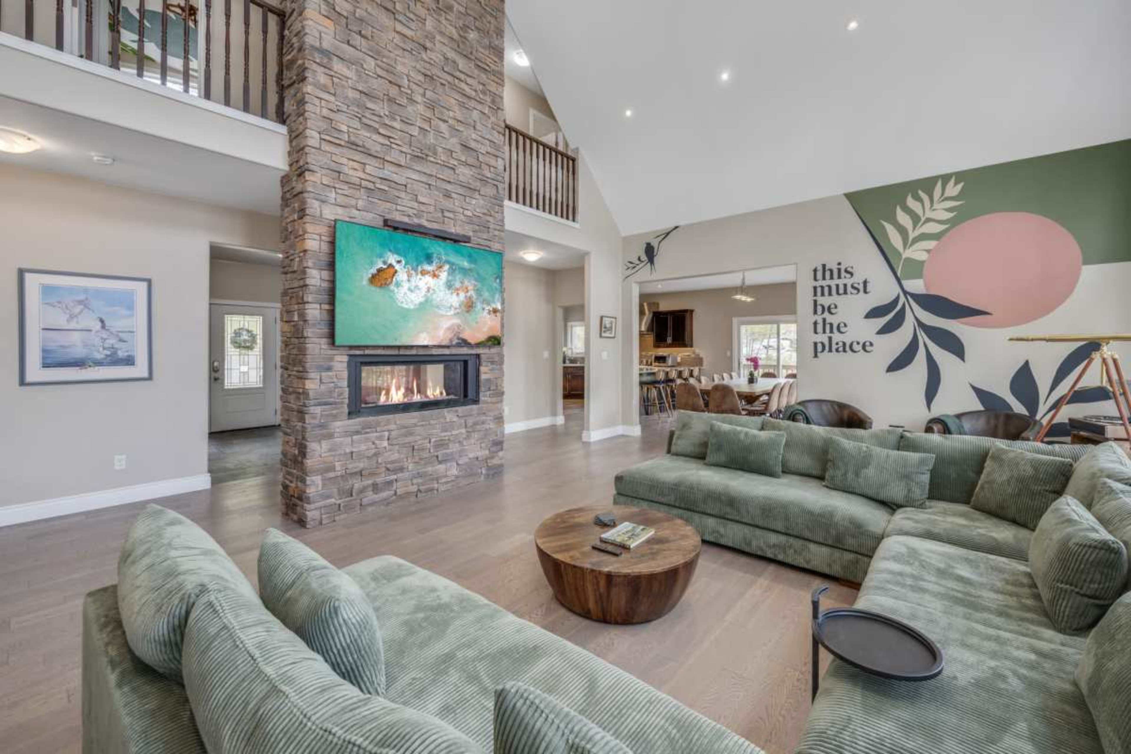 A spacious living area featuring a green sectional sofa, a stone fireplace with a mounted TV above it, and a wall mural with decorative elements.
