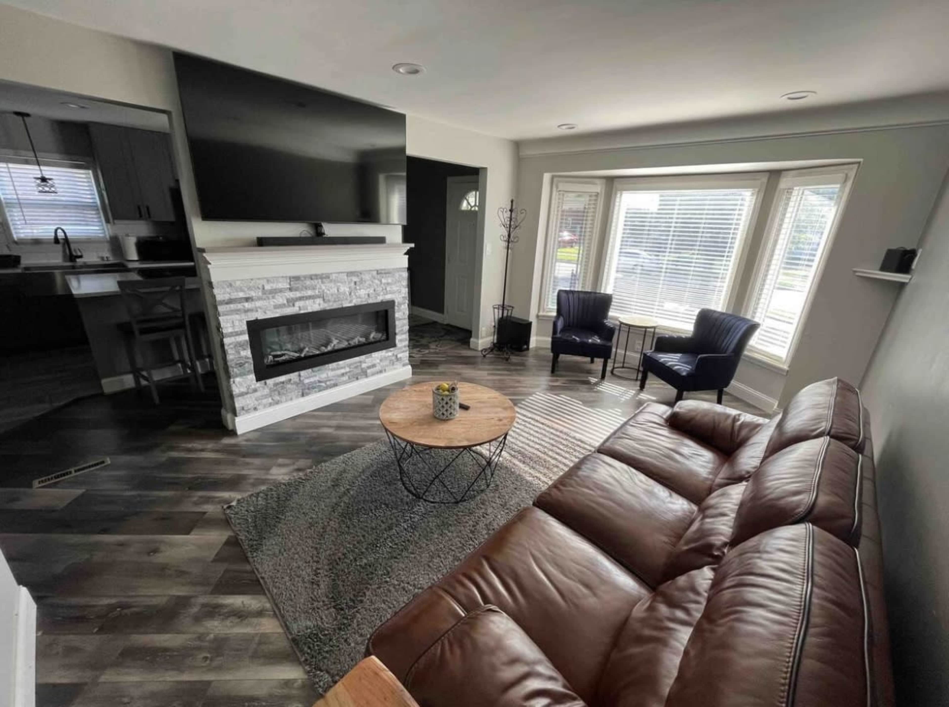 A modern living room featuring a brown leather sofa, a round wooden coffee table, two blue armchairs, and a wall-mounted television above a stone fireplace.