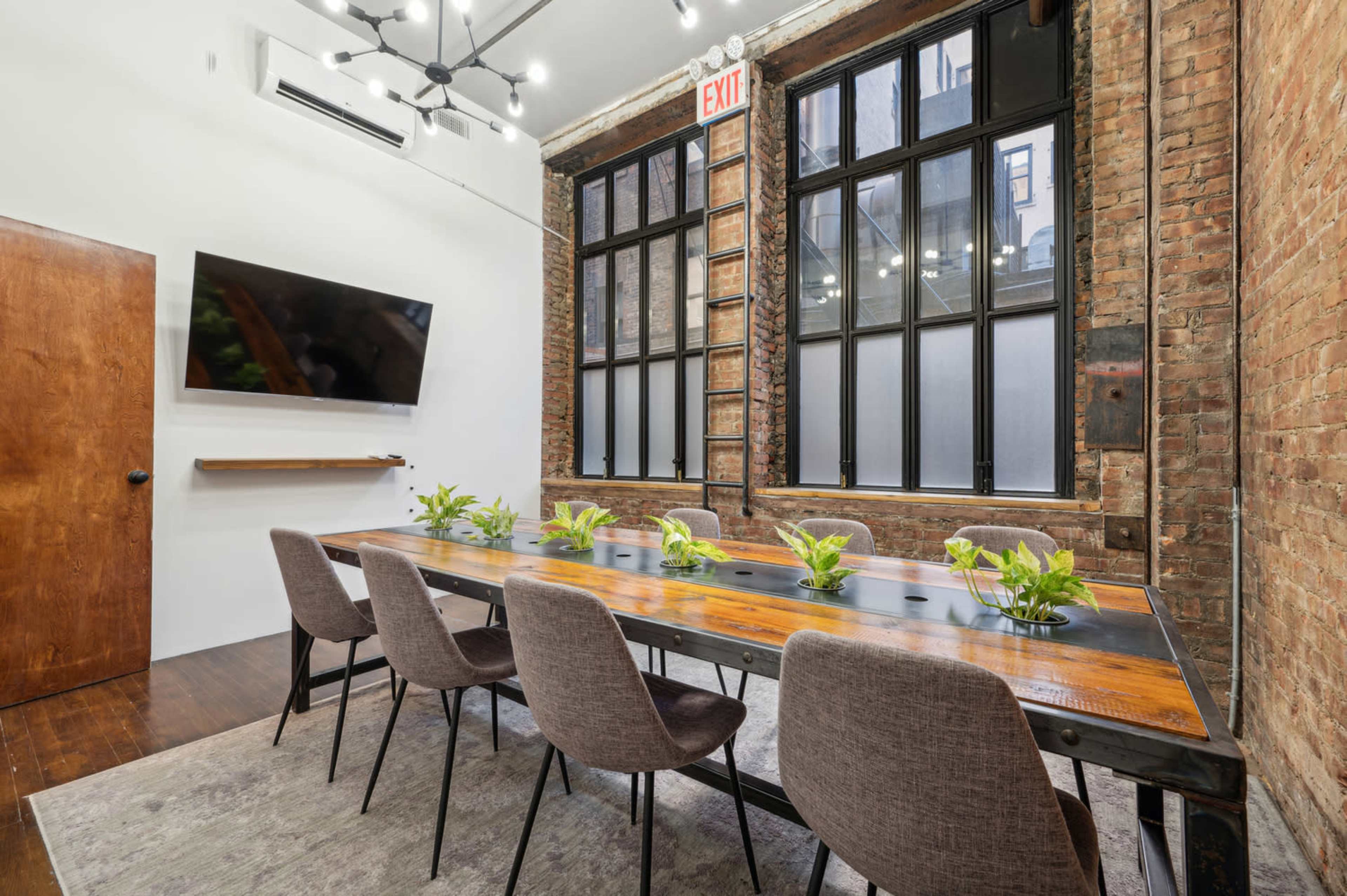 The image shows a modern conference room with a long wooden table surrounded by gray chairs, large windows, and a wall-mounted TV.