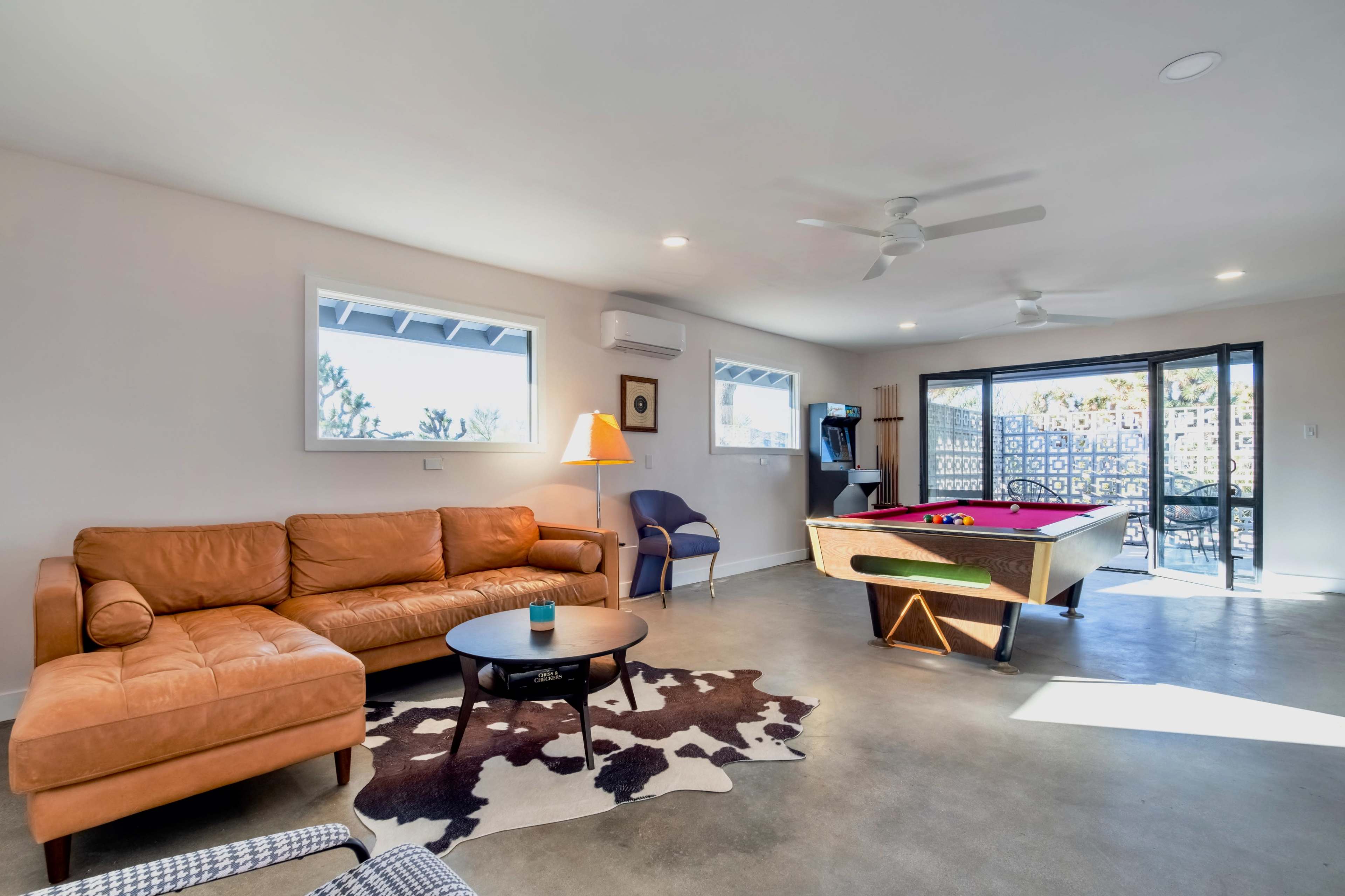 A spacious living room features a tan leather sofa, a round coffee table, and a pool table, with large windows allowing natural light to enter.