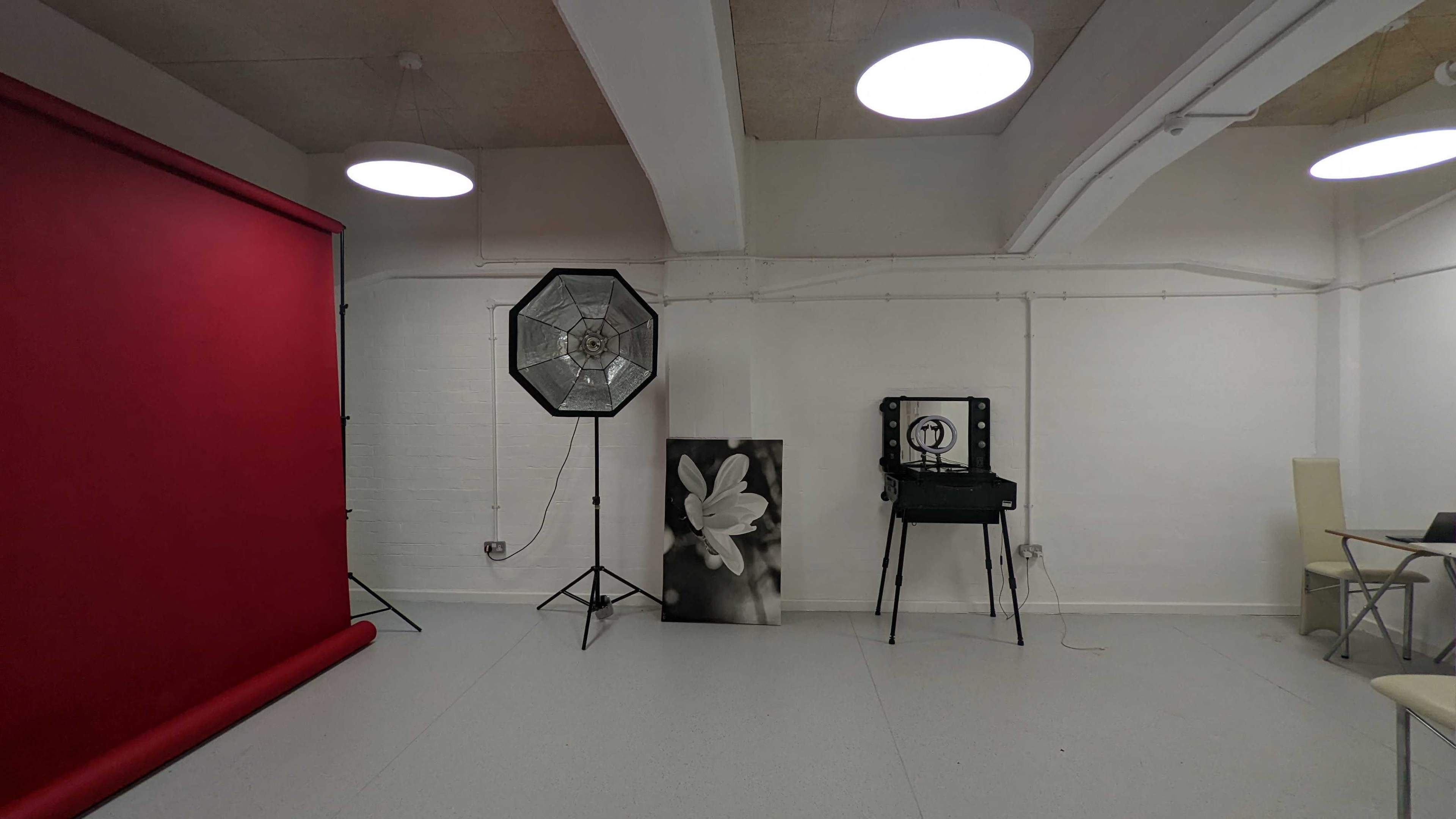 A photography studio featuring a red backdrop, a large softbox light, a black and white photograph of a flower, and an antique camera on a stand.
