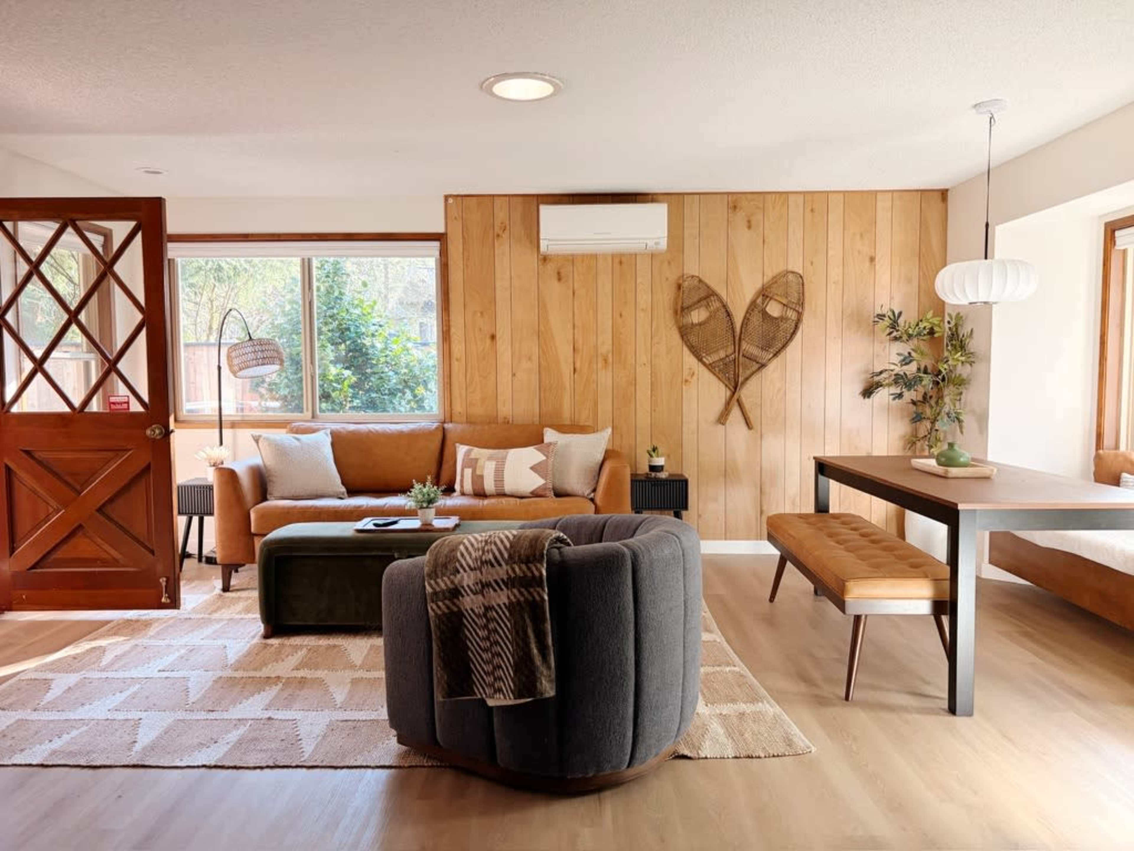 The image shows a cozy living room with a wooden accent wall, a sofa, a coffee table, and a dining area featuring a table and bench seating.