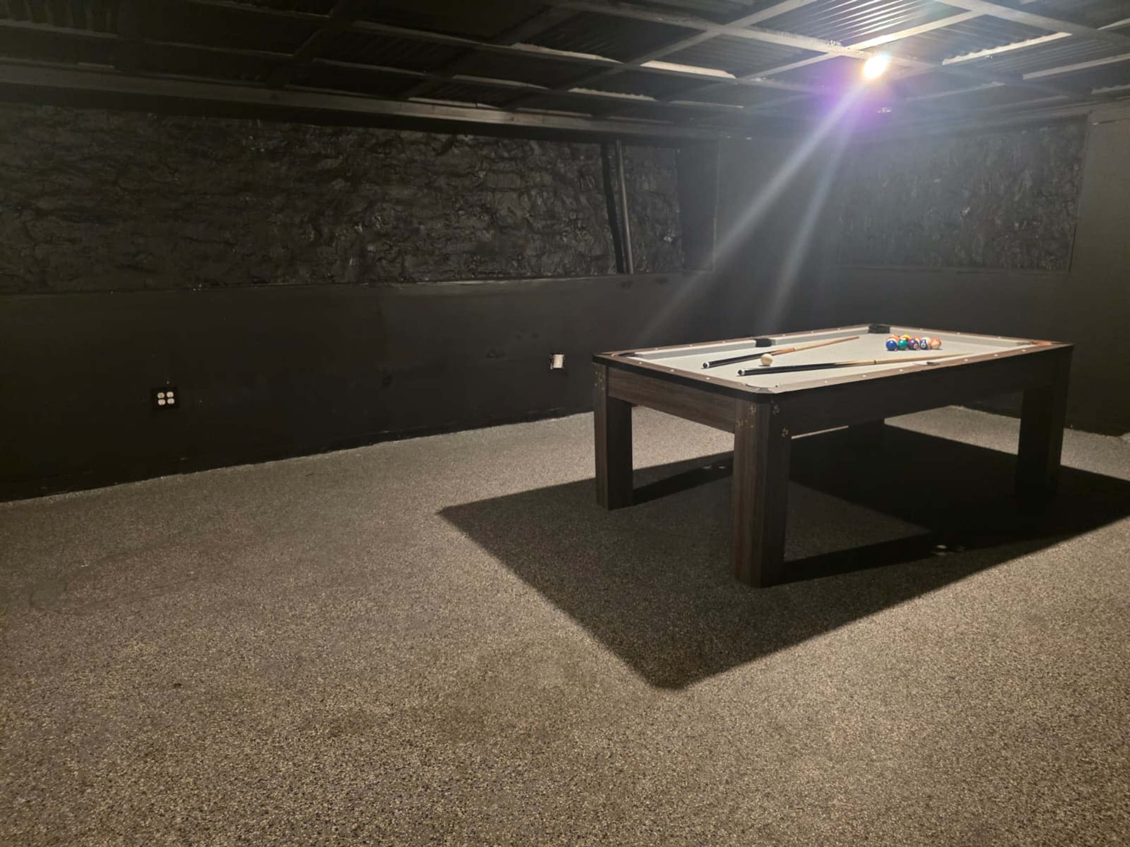 The image shows a dimly lit basement room with a pool table in the center and dark textured walls.
