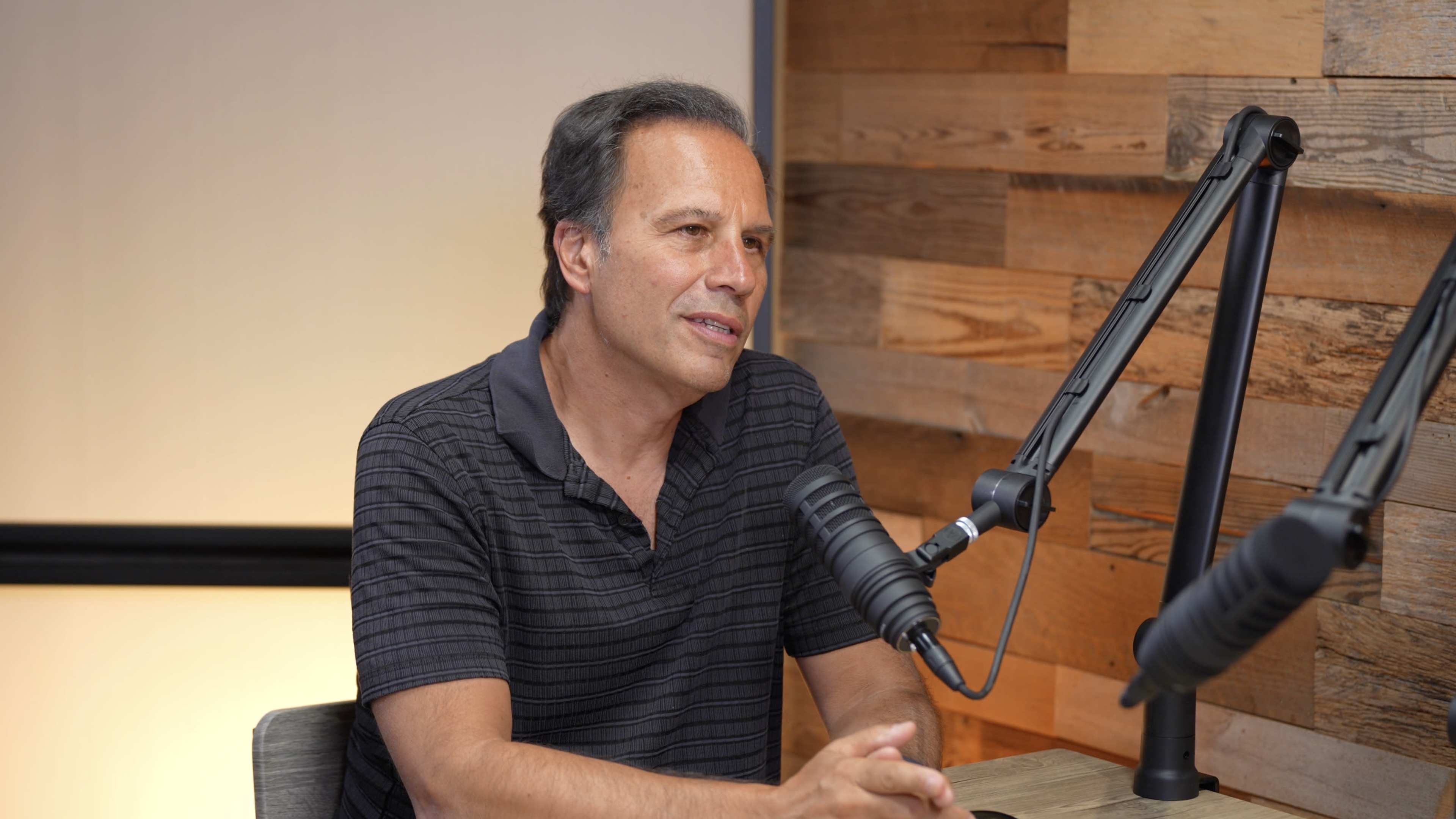A man in a black striped shirt sits at a wooden table speaking into a microphone in a room with wooden wall panels.