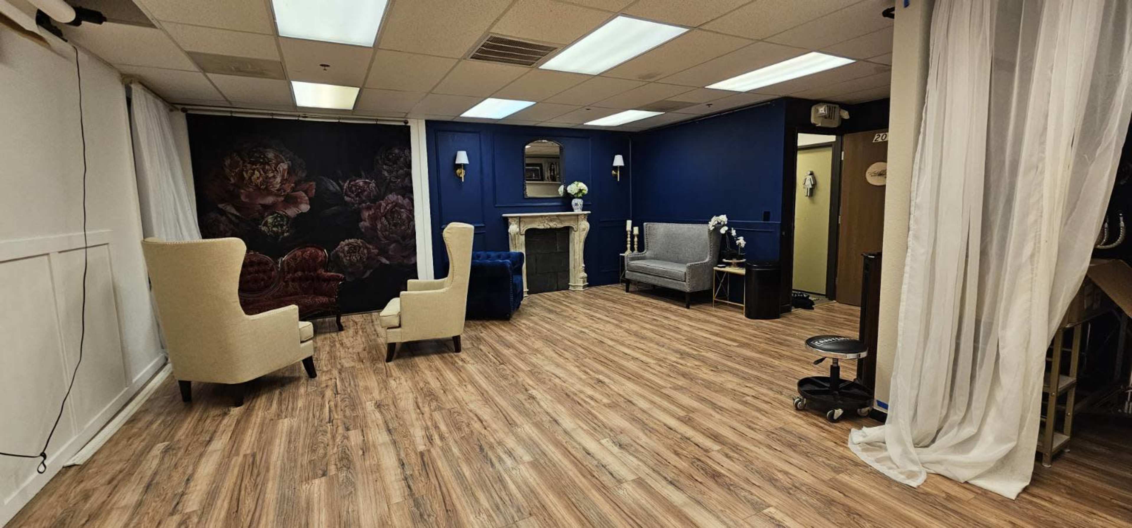 The image shows a spacious room with wooden flooring, two light-colored chairs, and a decorative wall featuring floral artwork.