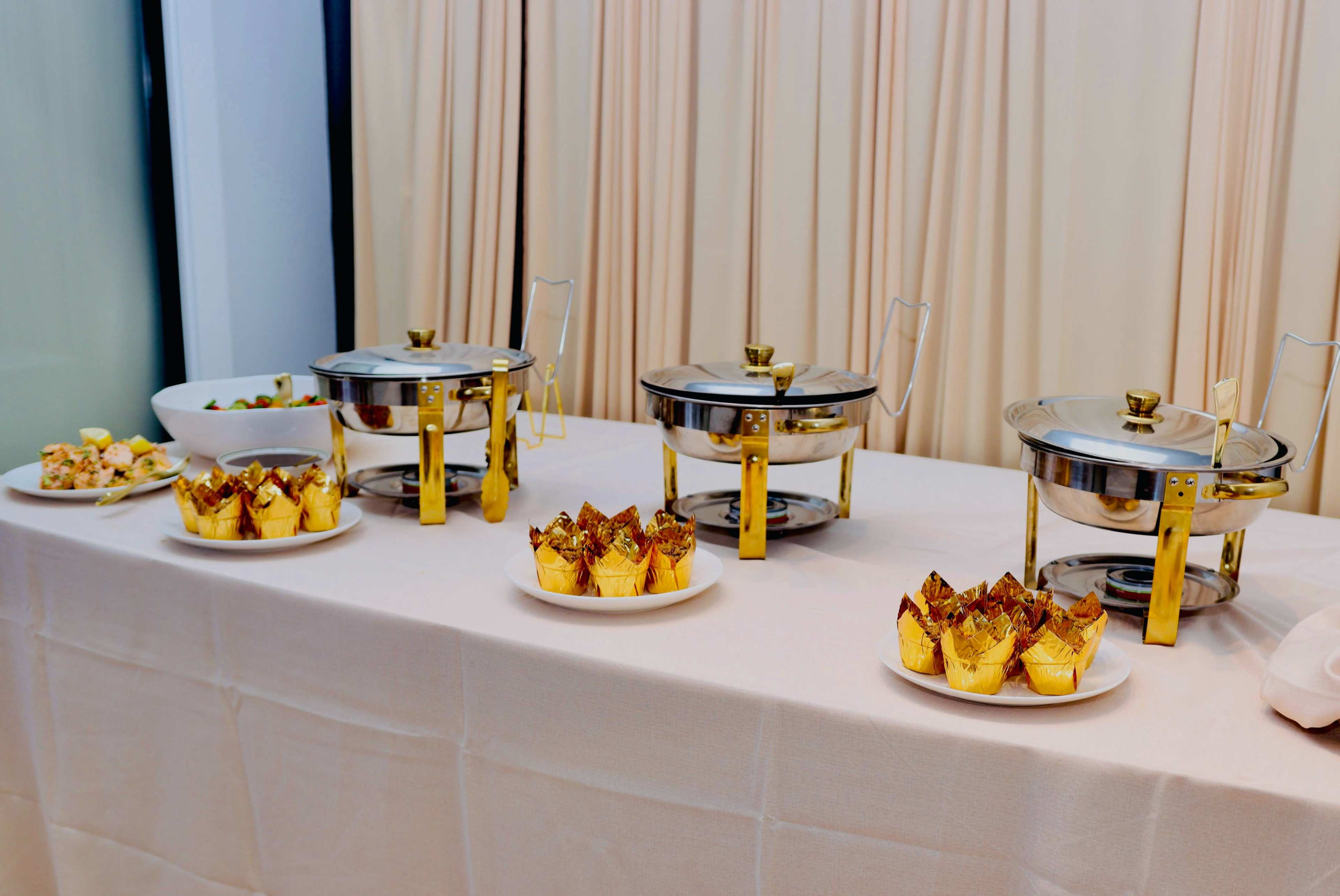 A table is set with chafing dishes and plates of dessert cupcakes, all arranged neatly with a backdrop of folded curtains.