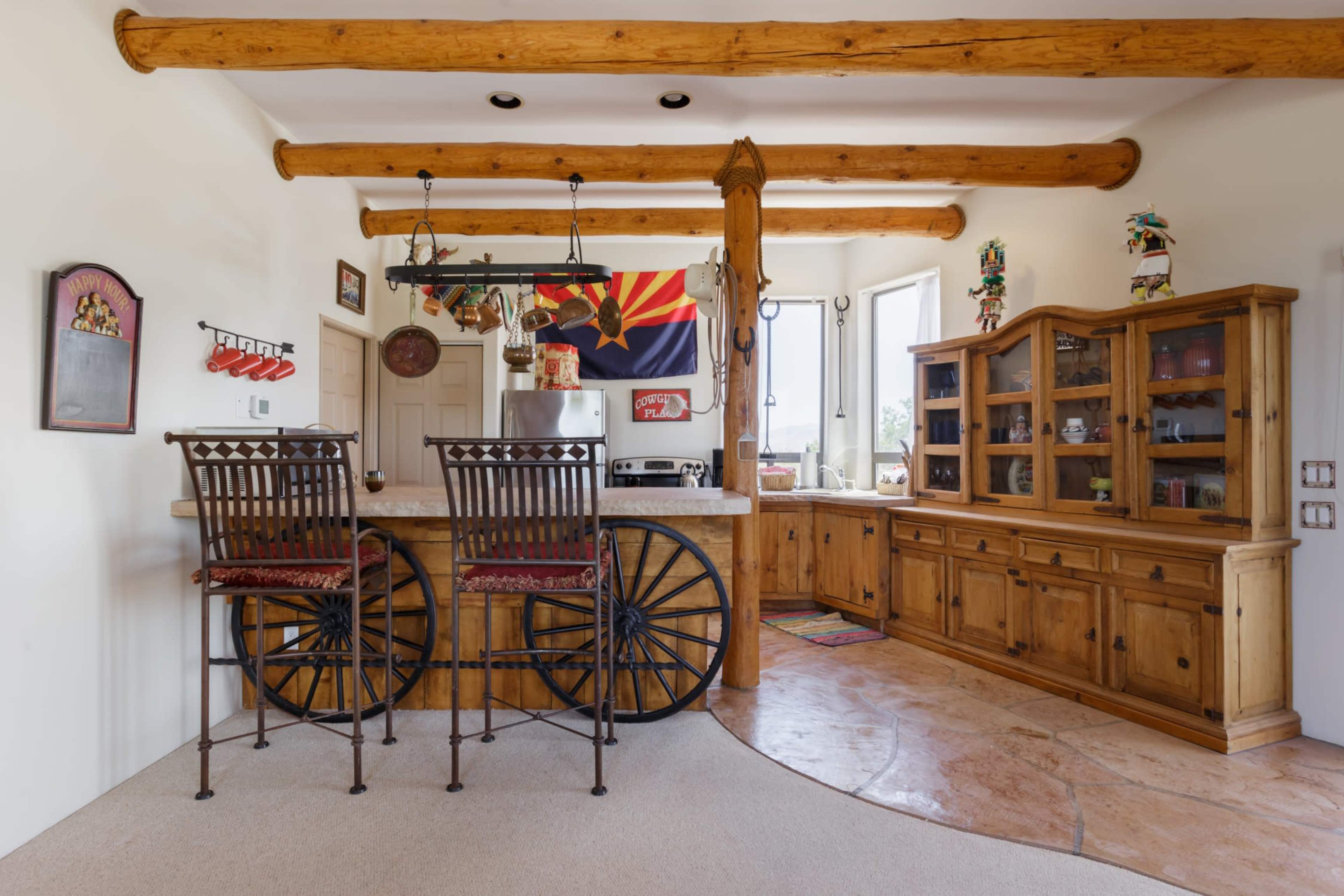 A rustic kitchen features a bar with high-backed chairs, wooden beams overhead, and a southwestern flag hanging near the window.