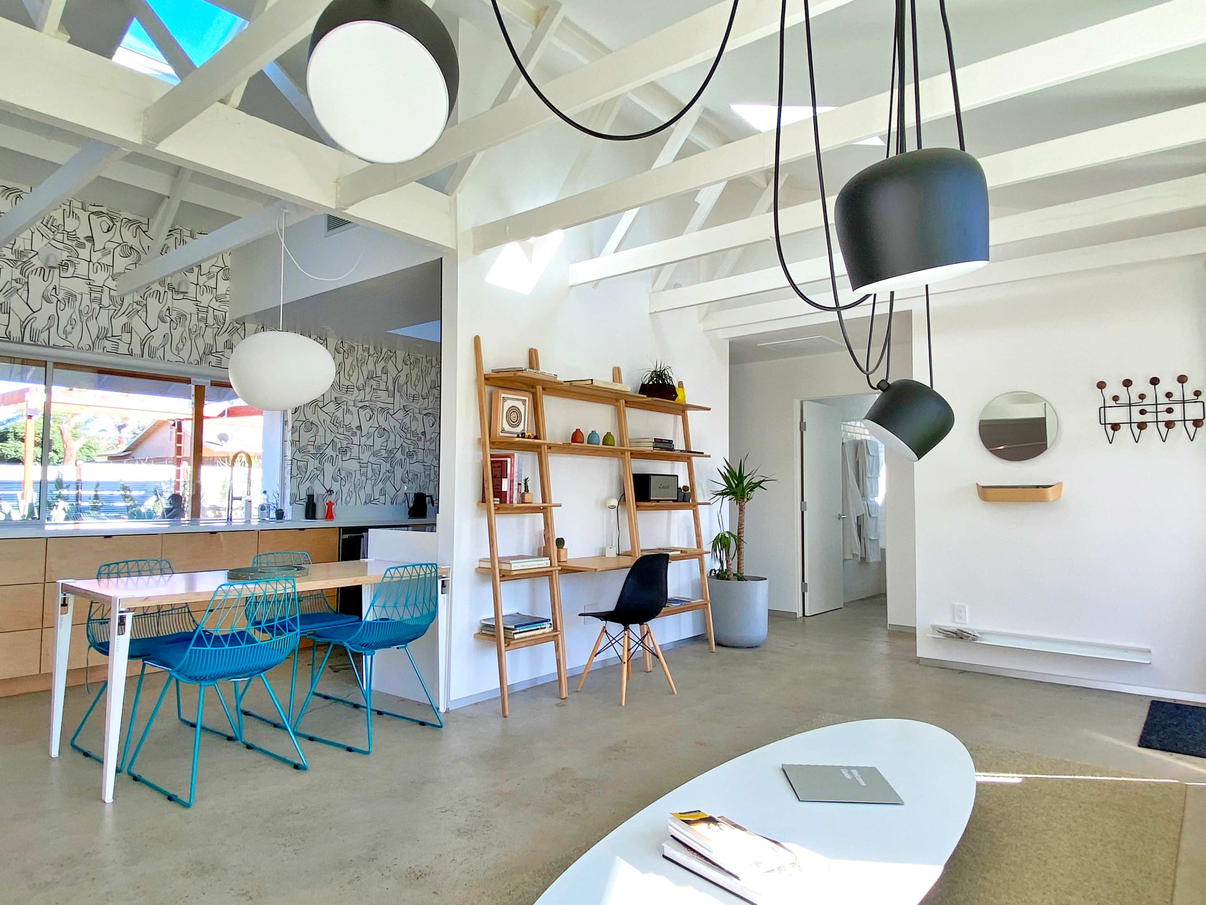 The image shows a modern, open-concept living space featuring a dining area with a wooden table and blue chairs, a bookshelf, and a minimalist decor.