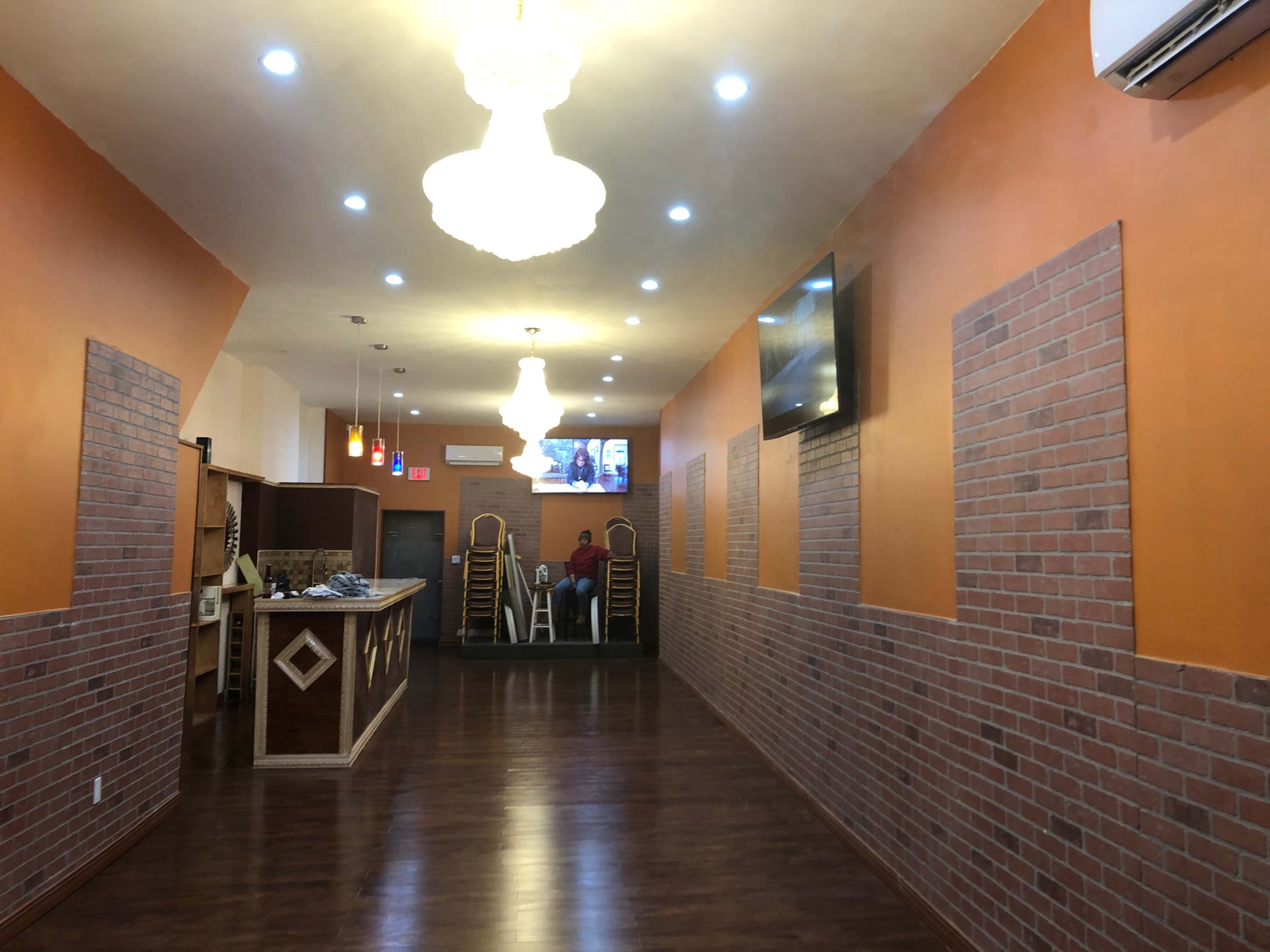 A hallway in a restaurant with brick-patterned walls, wooden flooring, and a reception area illuminated by chandeliers.