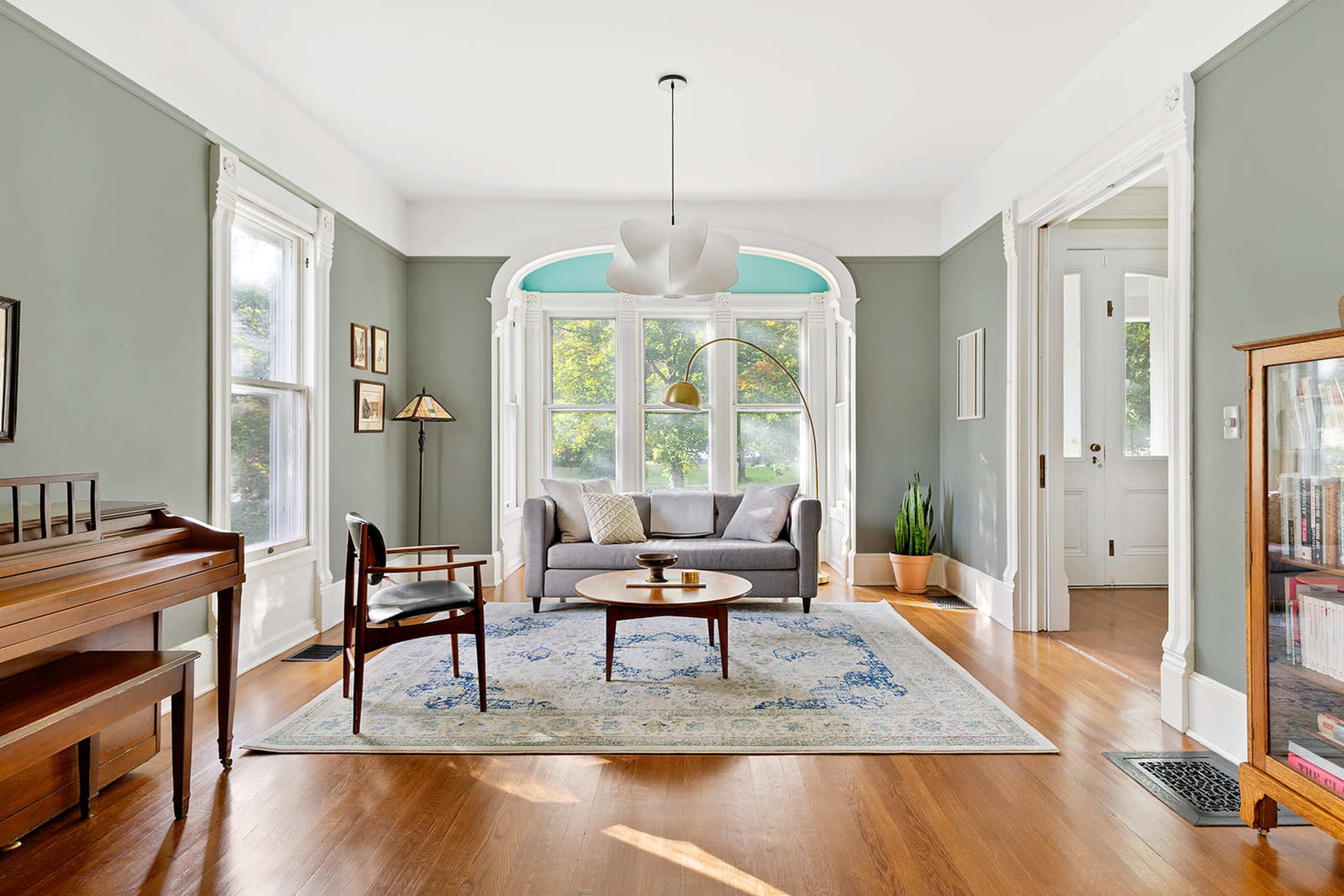 The image shows a well-lit living room featuring a gray sofa, a round coffee table, a piano, and large windows with green accents.