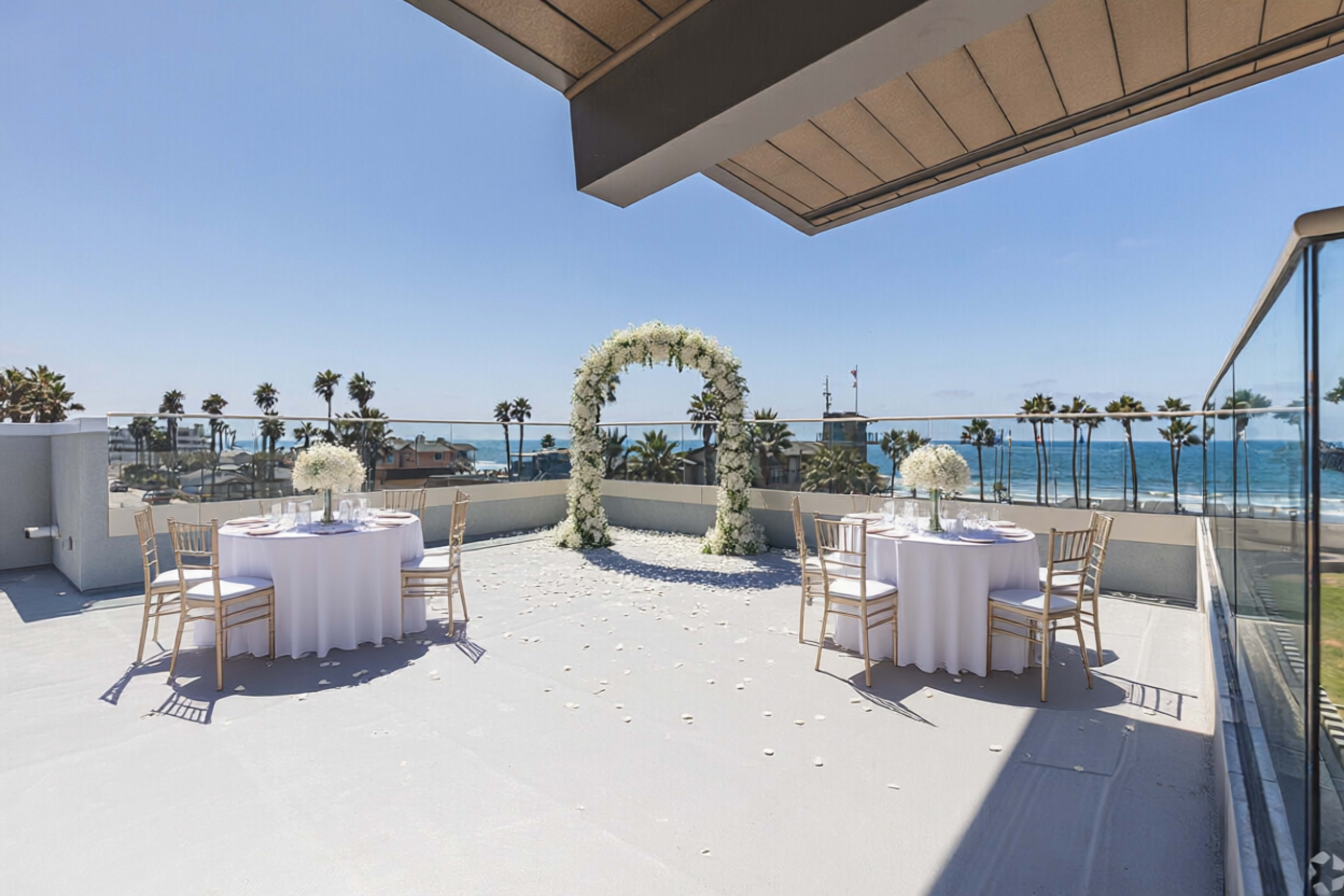 A rooftop venue features two round tables with white tablecloths set for an event, and an ornate floral arch is positioned in front of a view of the ocean and palm trees.