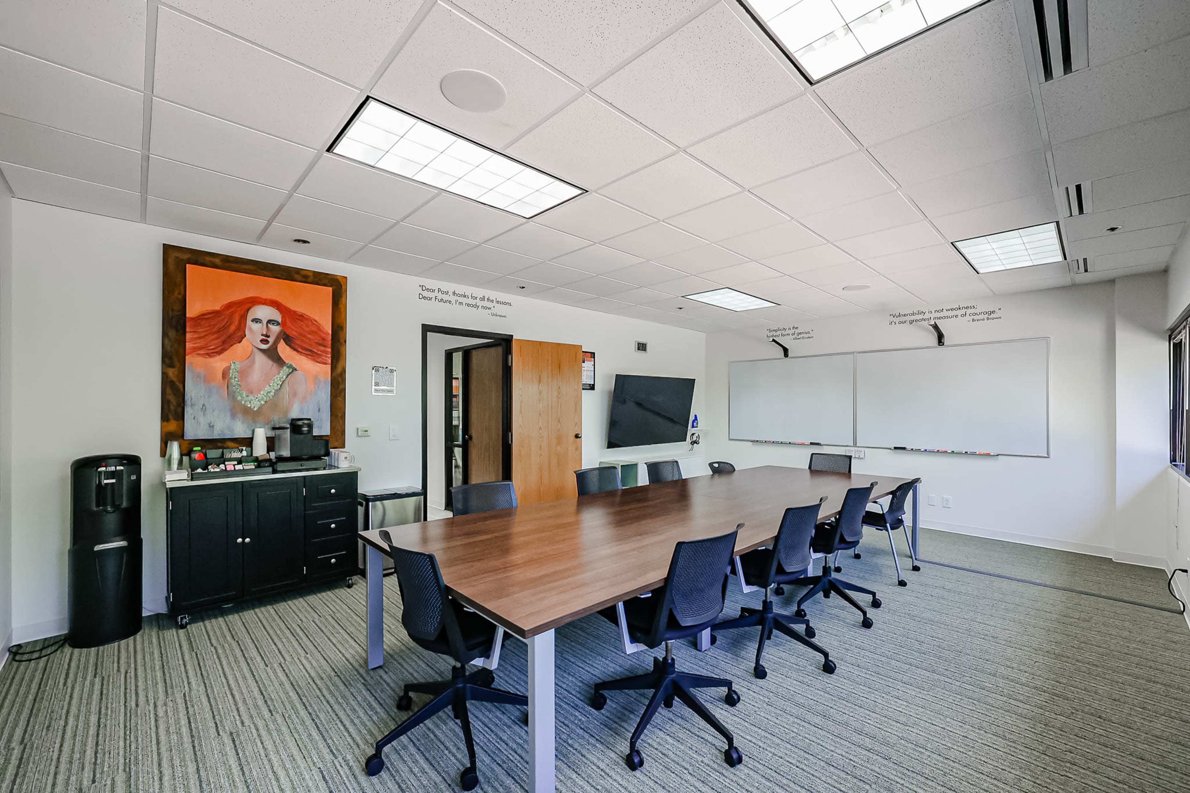 A conference room with a long wooden table surrounded by ergonomic chairs, a wall-mounted television, and a large artwork featuring a woman on one wall.