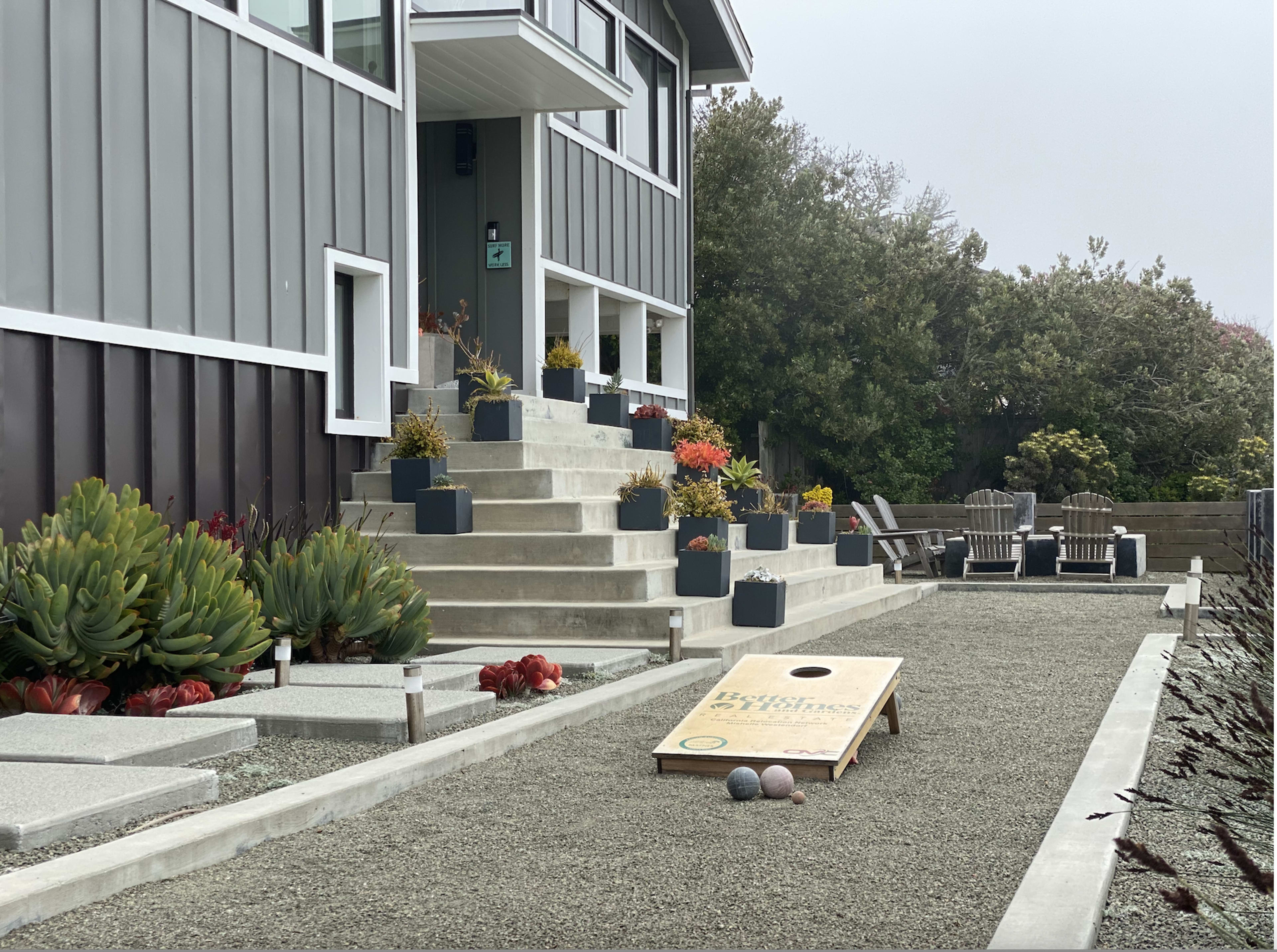 A gravel pathway leads to a modern house with multiple steps, bordered by planters and featuring a cornhole game set up in the yard.