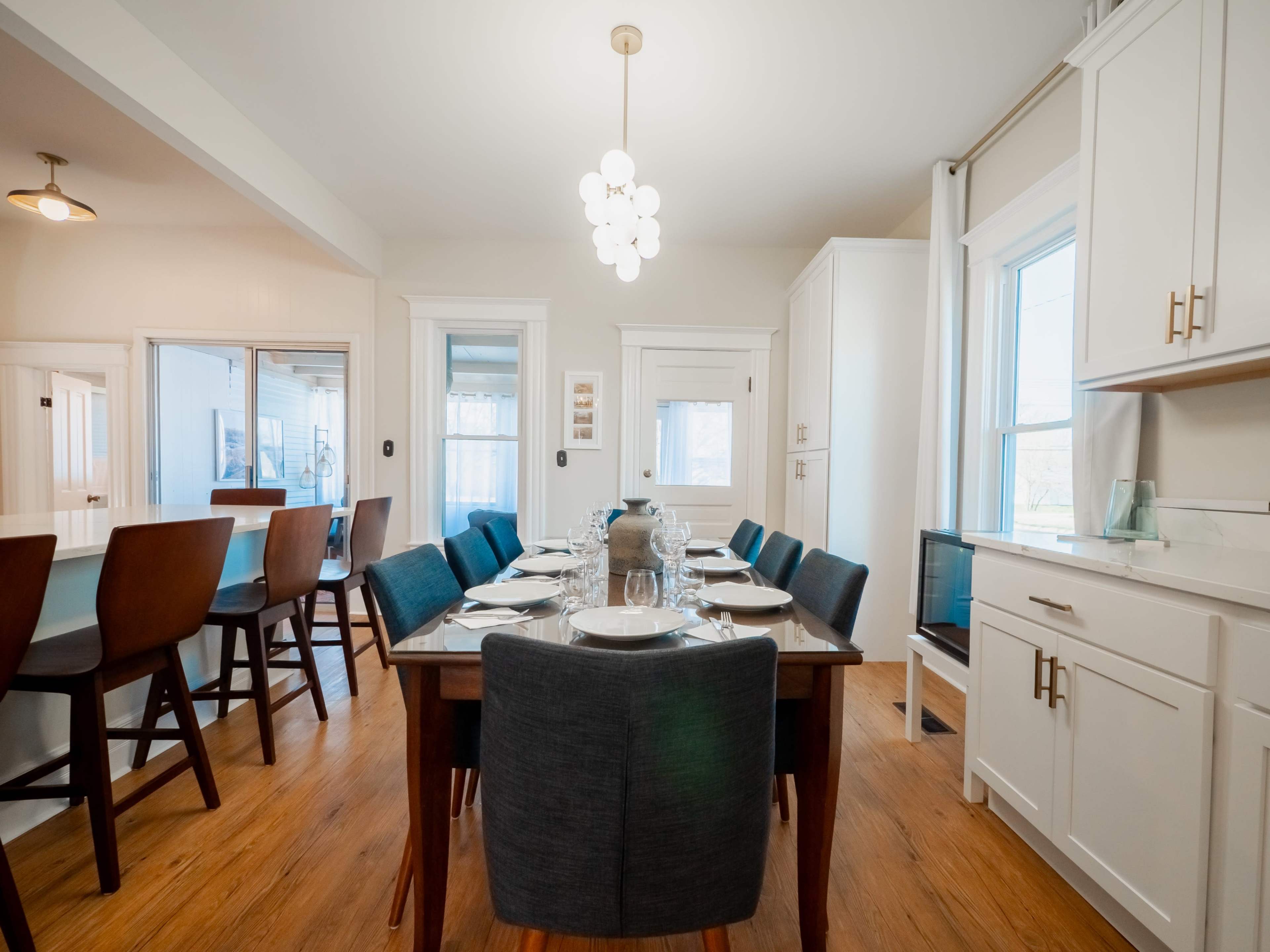A dining room features a long table set with plates and glasses, surrounded by upholstered chairs, with large windows allowing natural light to enter.