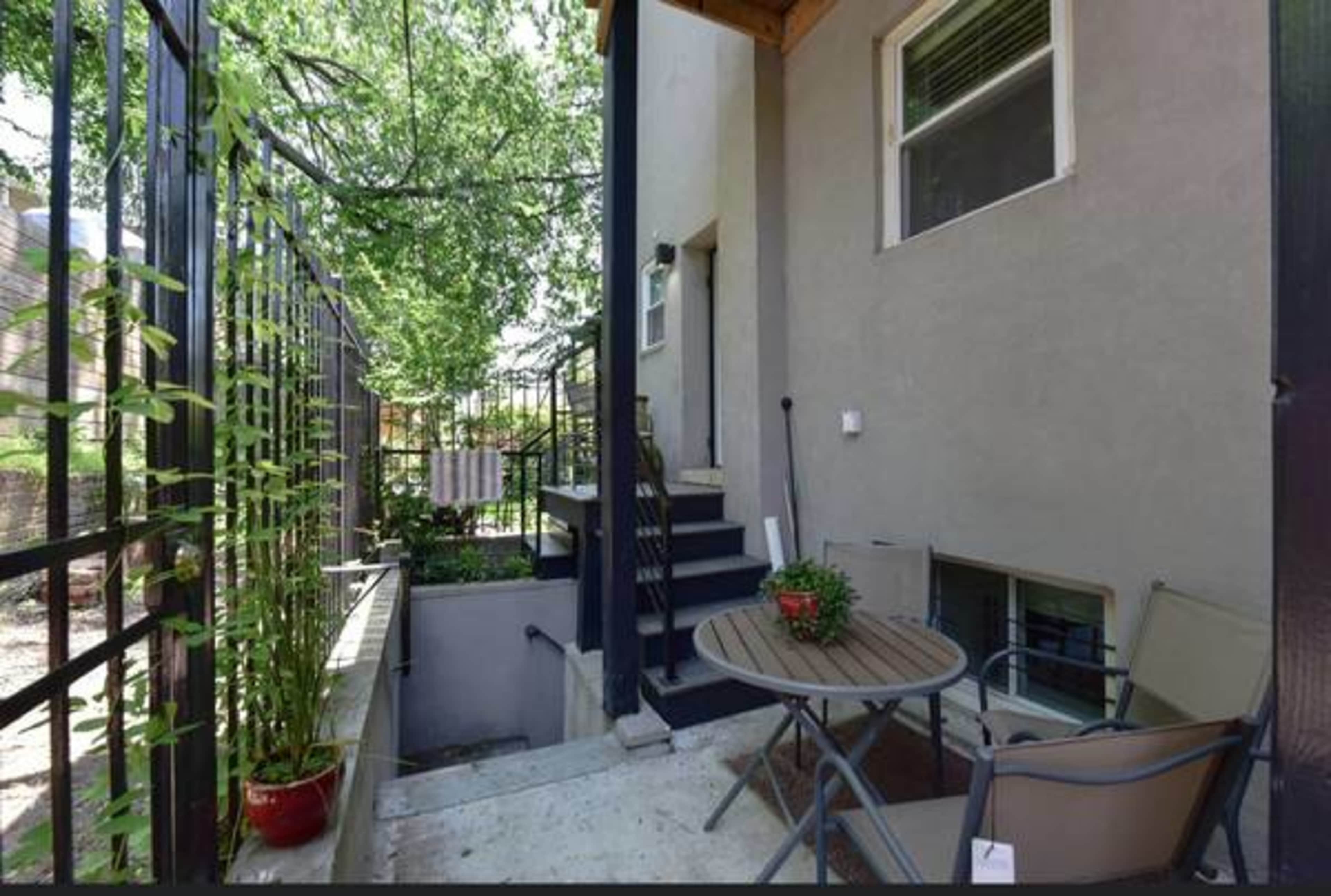 A small outdoor patio area featuring a table and chairs, surrounded by greenery and with stairs leading to an entrance.