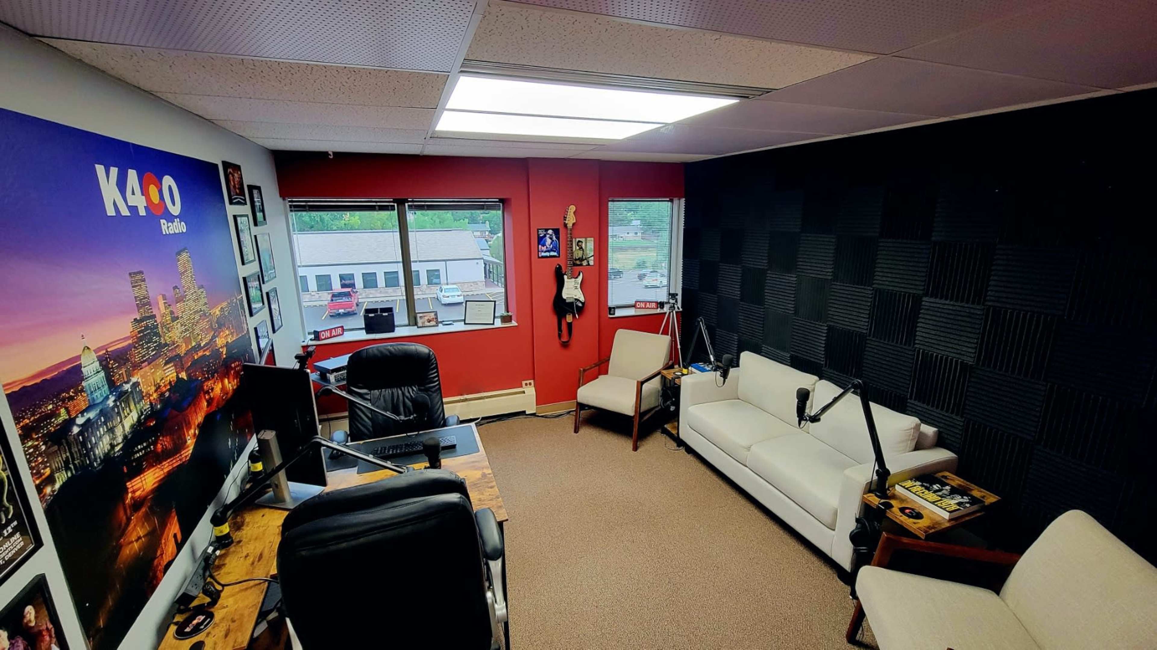 The image shows a small radio studio with a desk, chair, white sofa, and soundproof black wall panels.