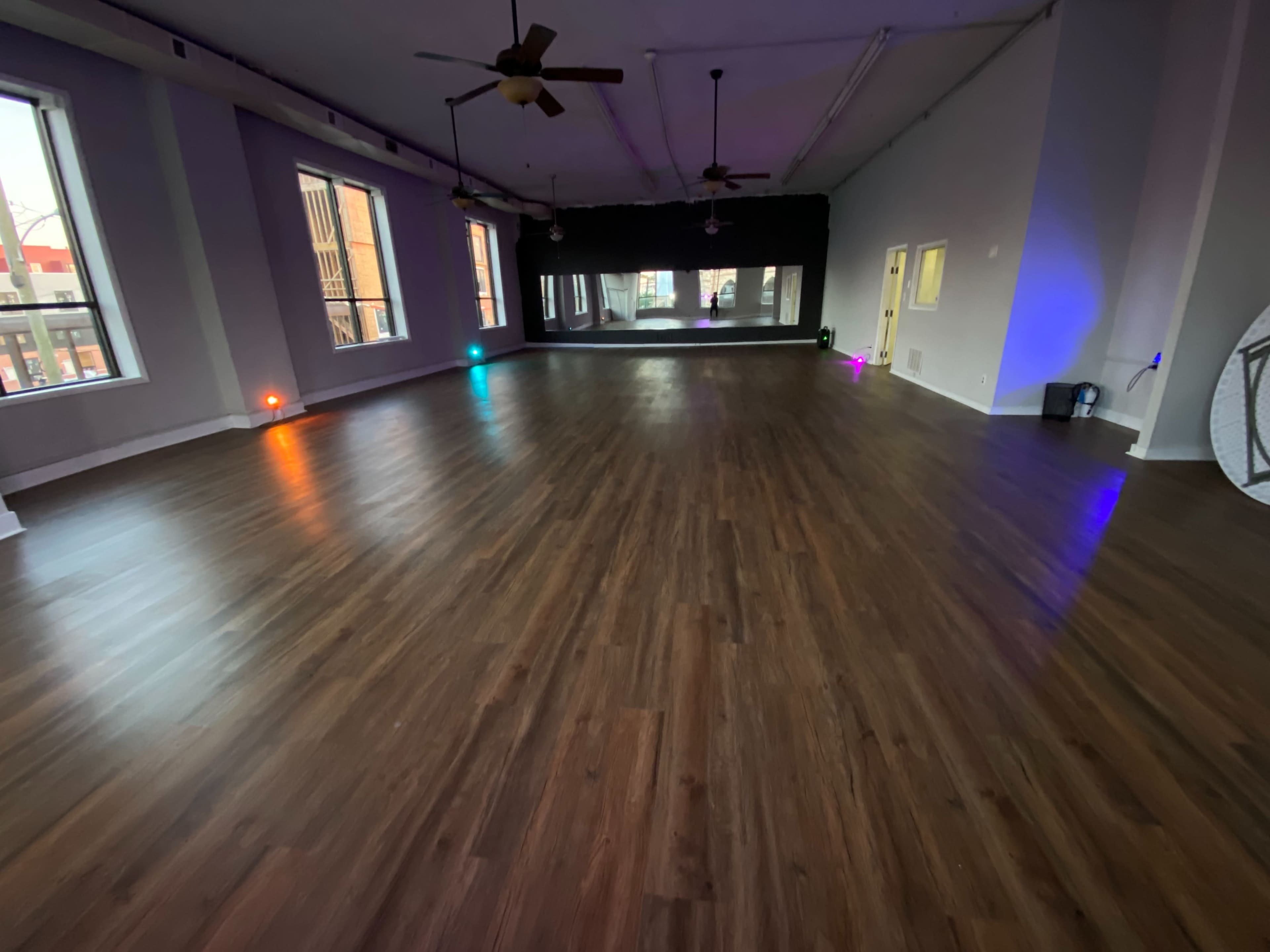 A spacious, empty room with a wooden floor, large windows, and colored lights at the corners.