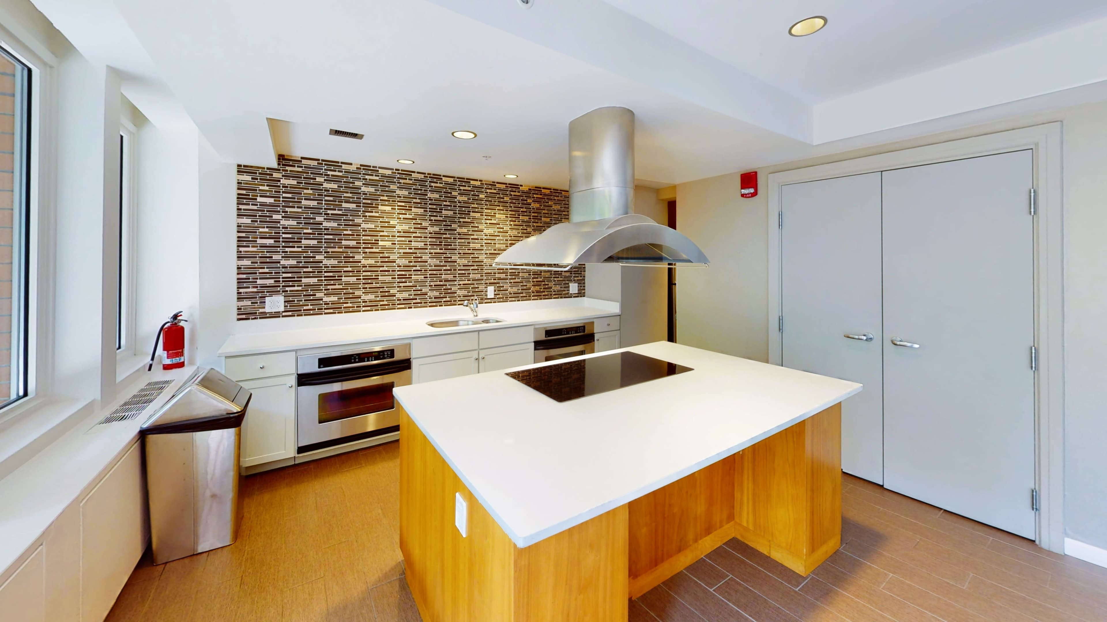 The image shows a modern kitchen featuring a large island with a cooktop, stainless steel appliances, and a mosaic tile backsplash.