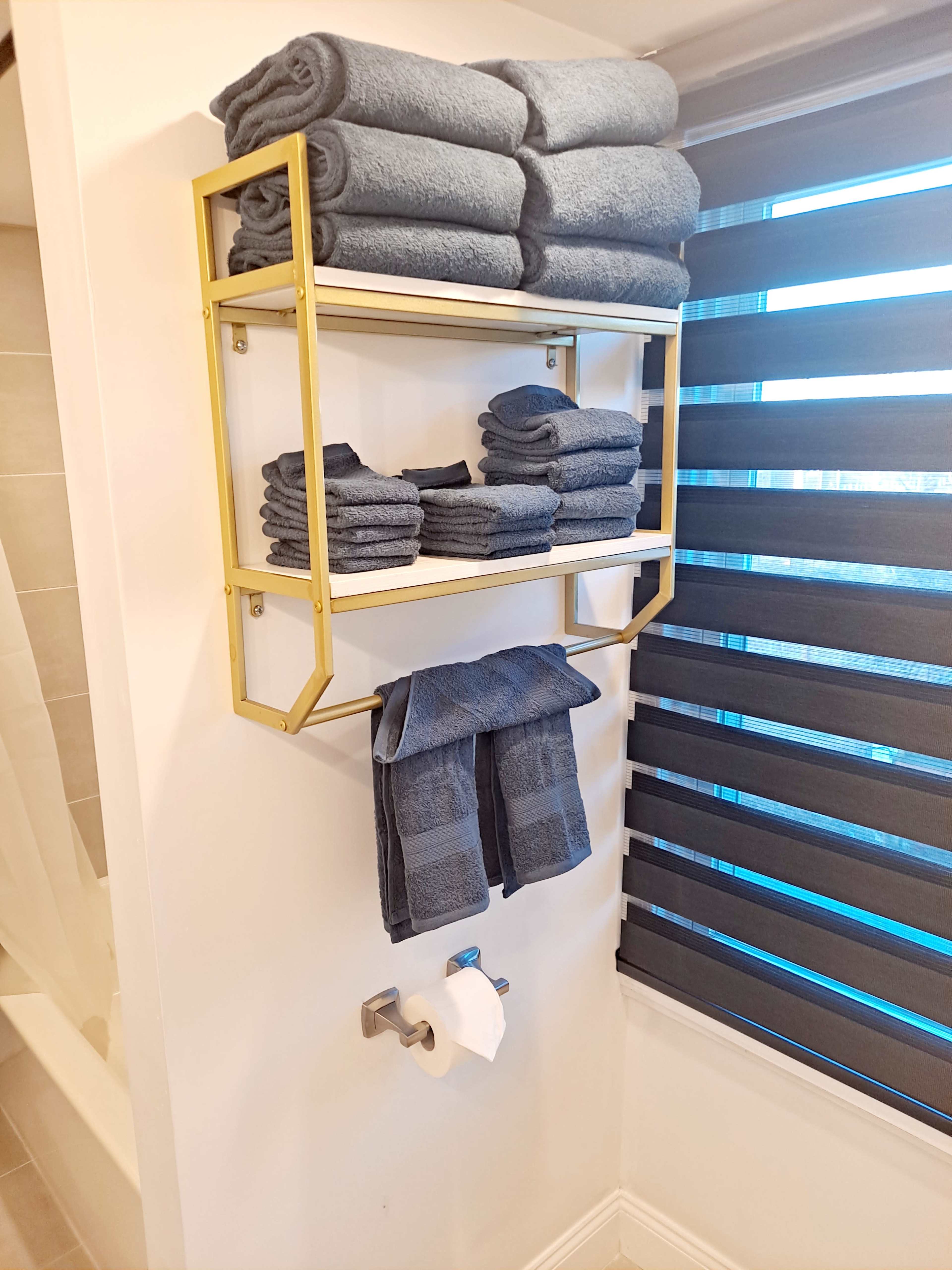 A gold-framed shelving unit displays neatly stacked dark blue towels next to a roll of toilet paper in a bathroom setting.