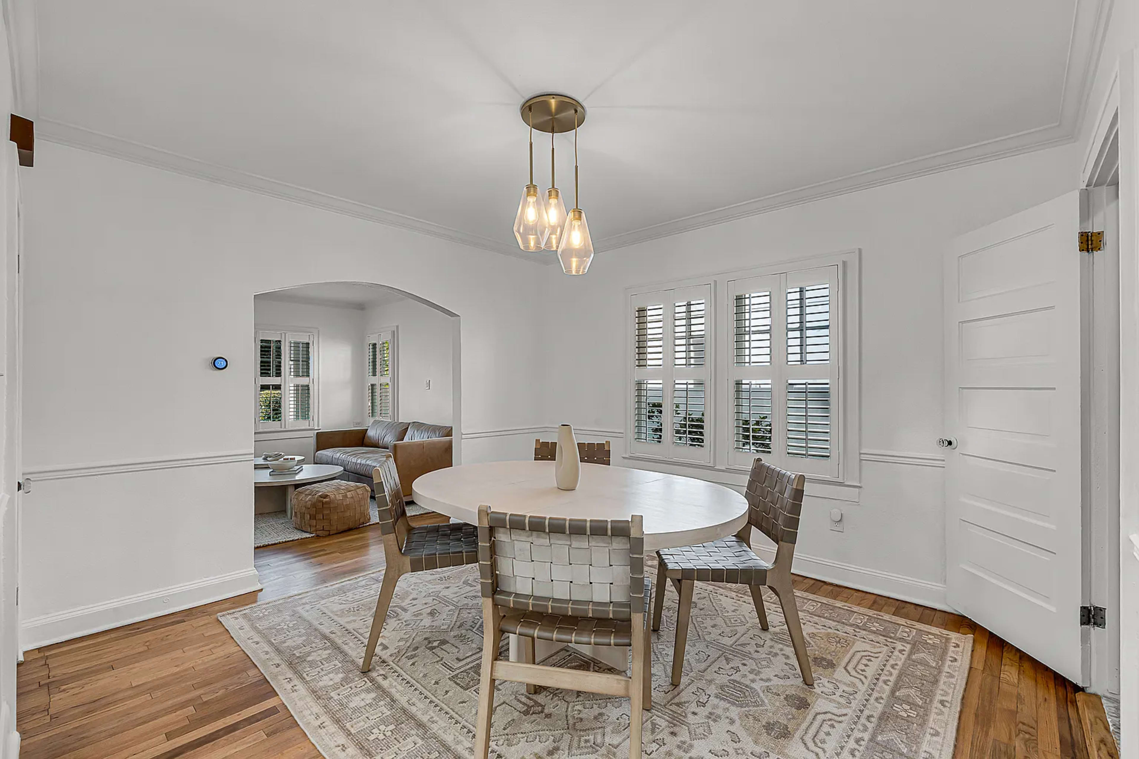 A round dining table with six chairs is set in a bright room, adjacent to a living area visible through an archway.