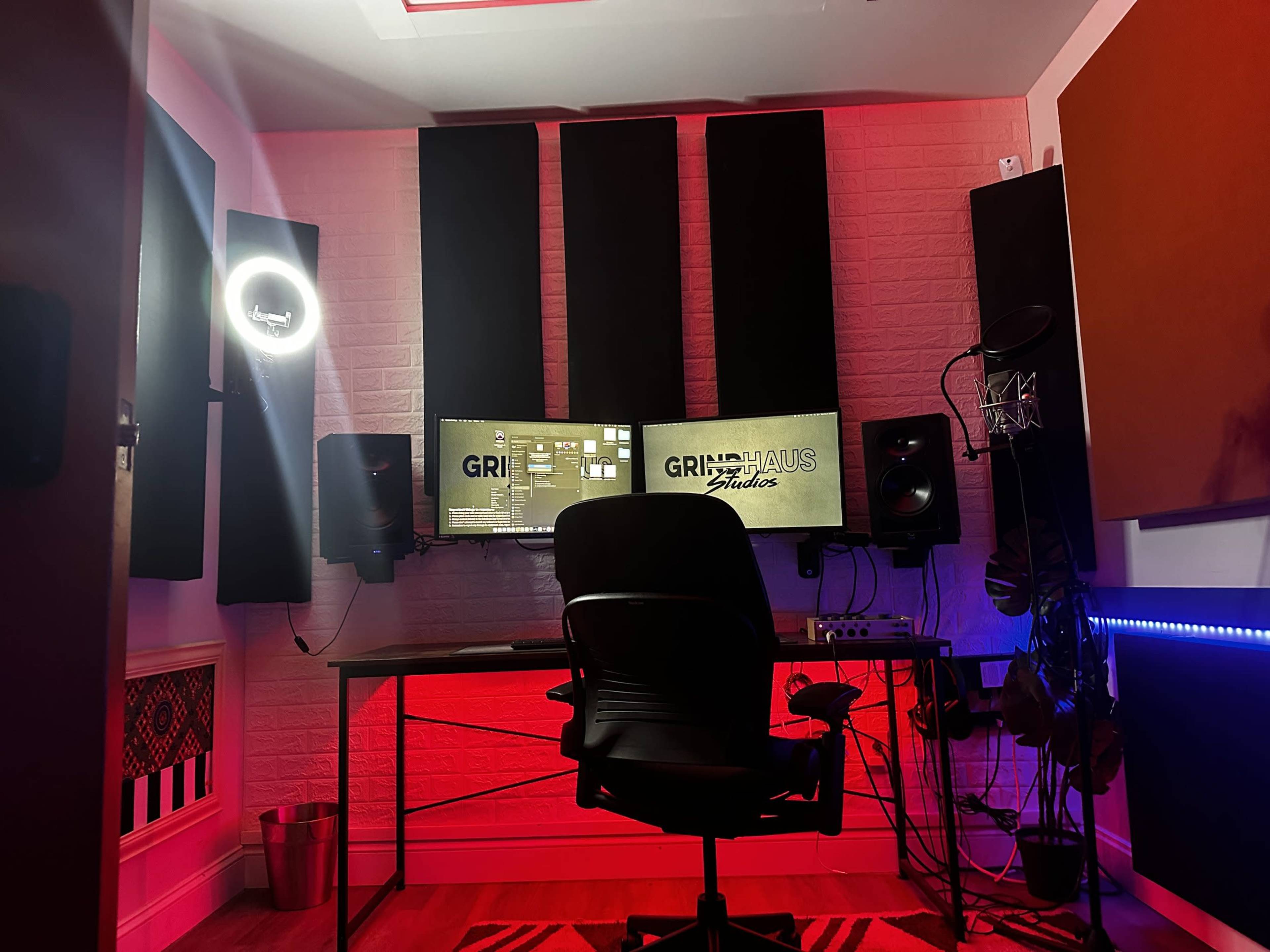 A music studio setup features a desk with a chair, dual speakers, and a large monitor displaying the logo "GRIMHAUS Studios," all illuminated with colorful lights.