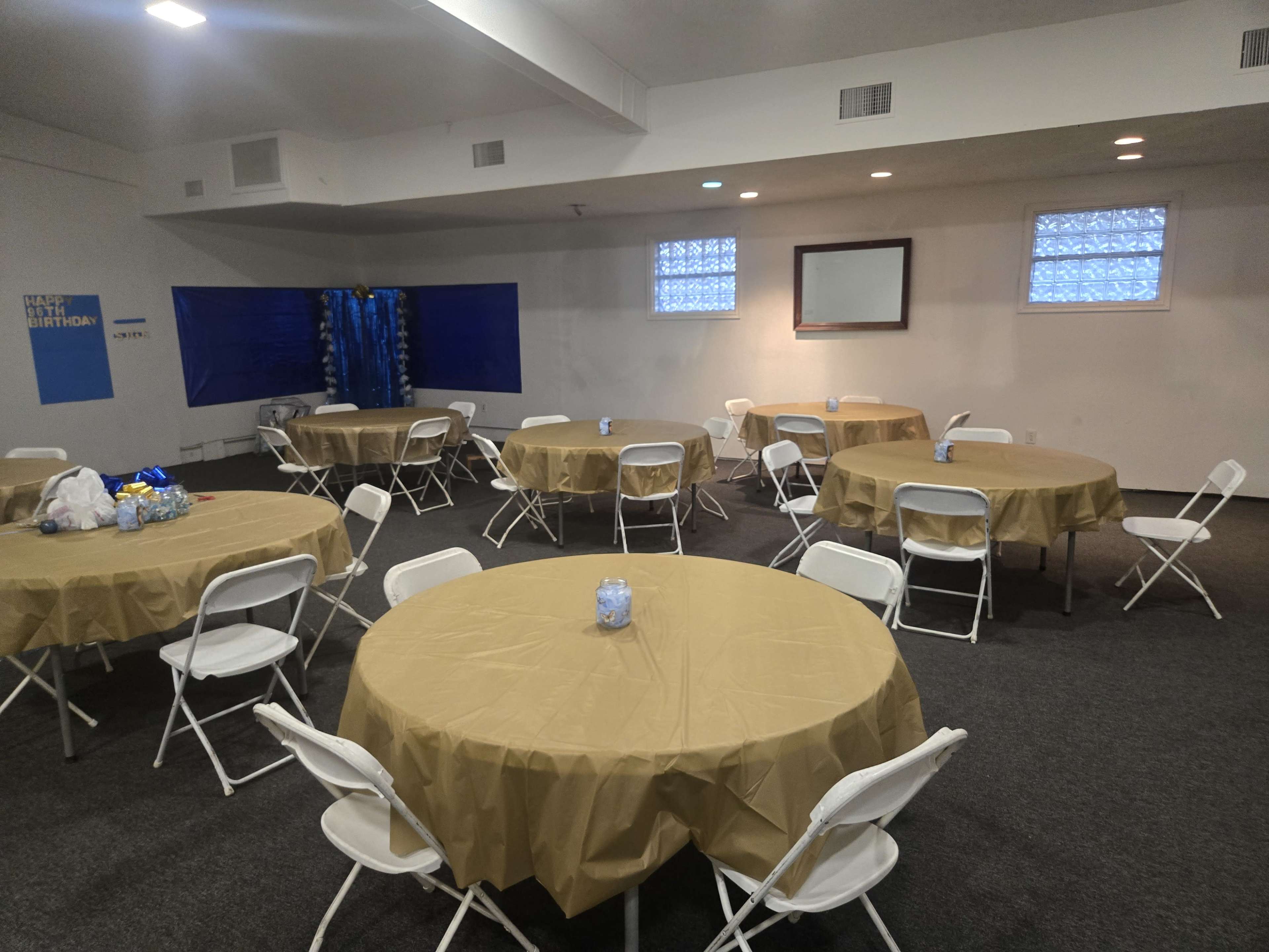 The image shows a room set up for an event, featuring several round tables covered with beige cloths and surrounded by white folding chairs.