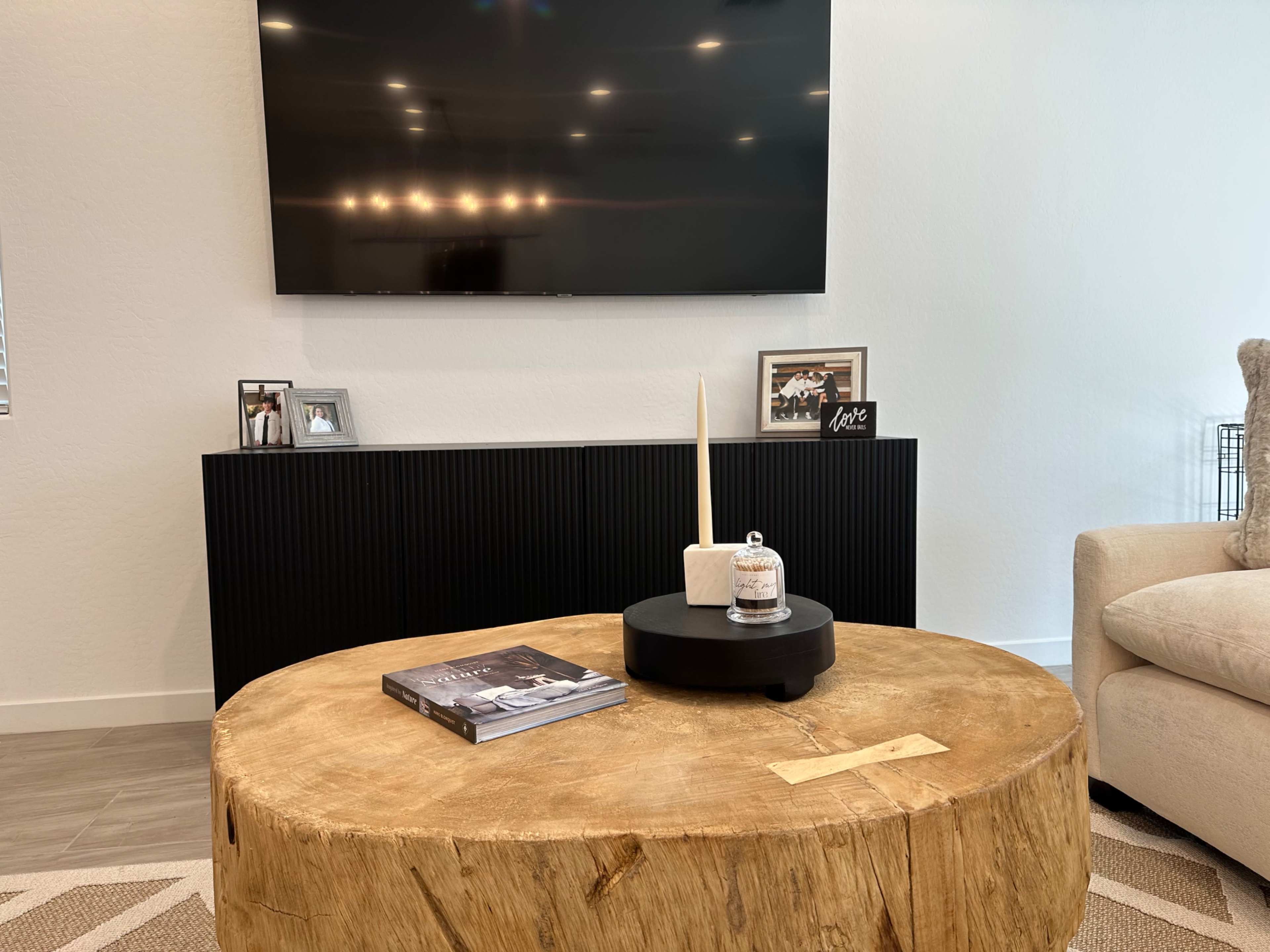A living room scene with a large wooden coffee table at the center, a modern black entertainment unit with a TV mounted above it, and decorative items on the table.