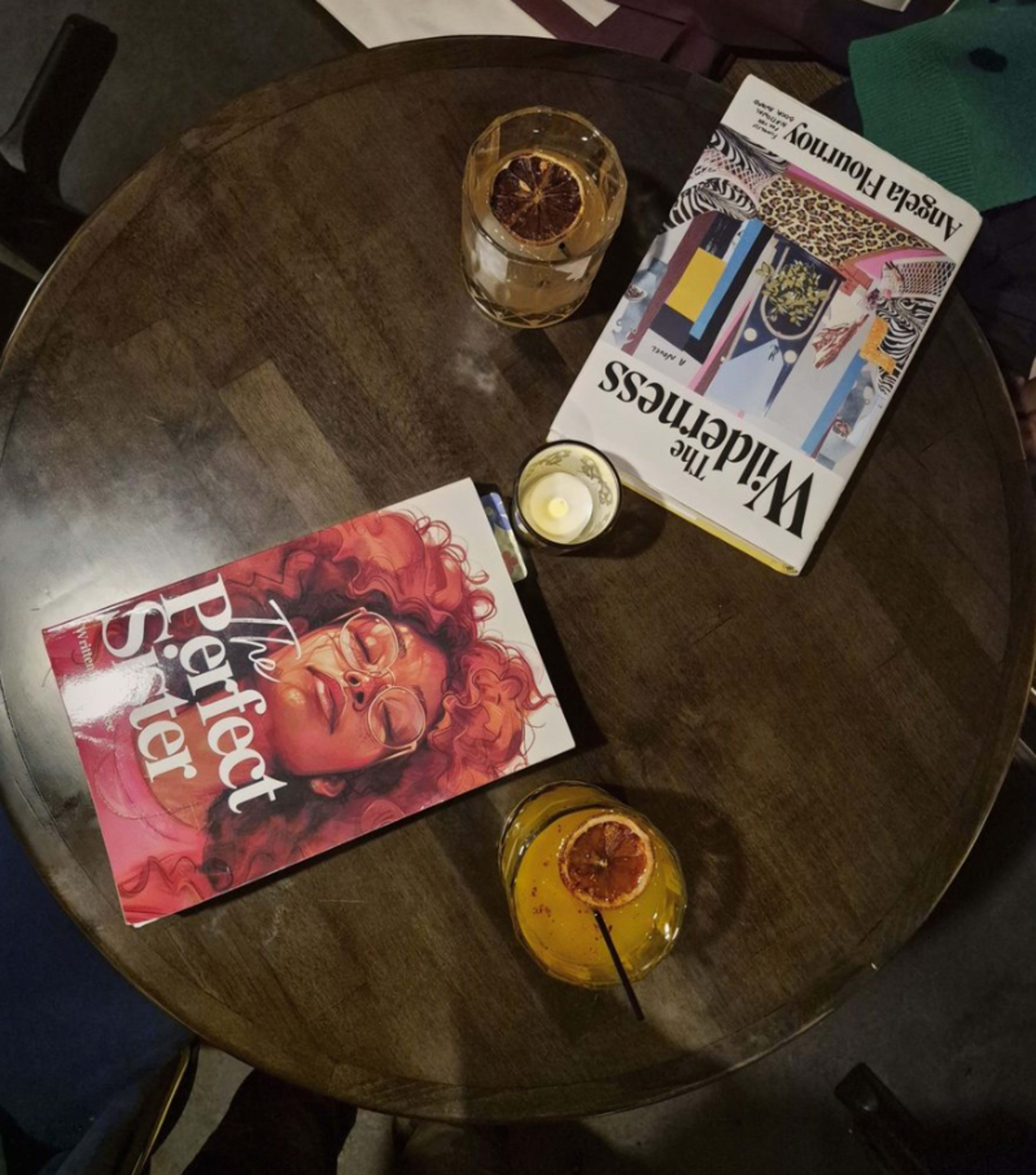 A round wooden table holds two books and two drinks, one featuring a large cover illustration of a woman and the other displaying colorful fashion designs.
