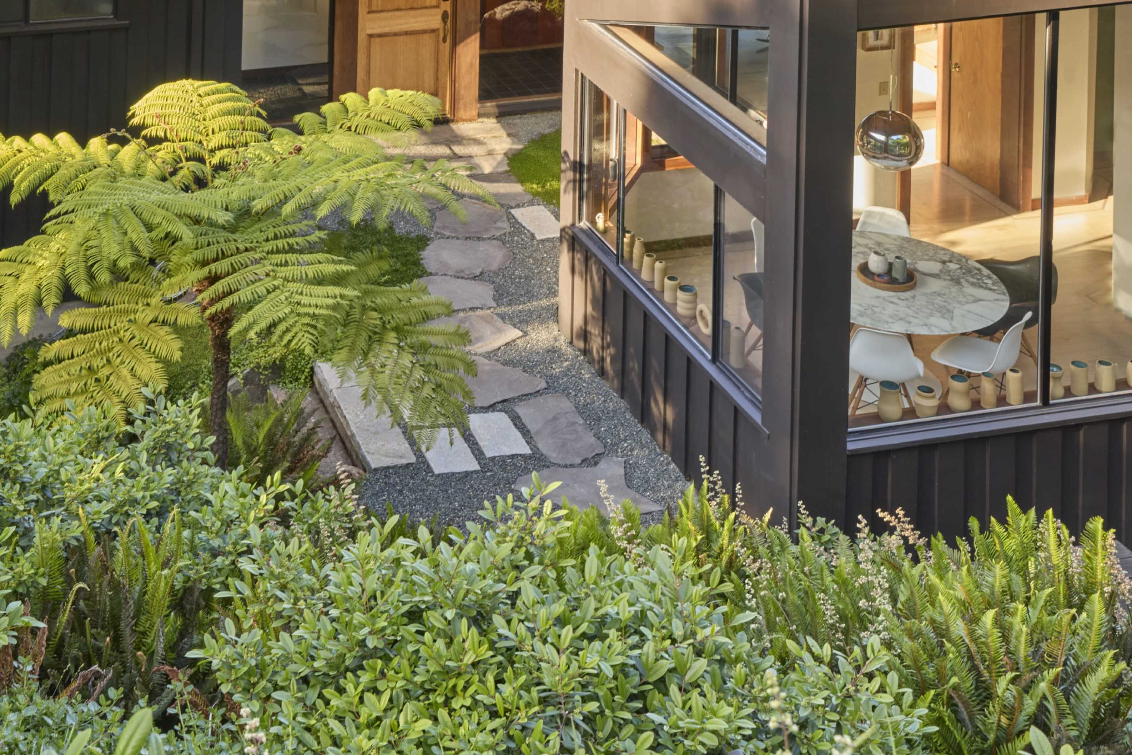The image shows a landscaped pathway made of stones and surrounded by greenery, leading to a modern building with large windows.