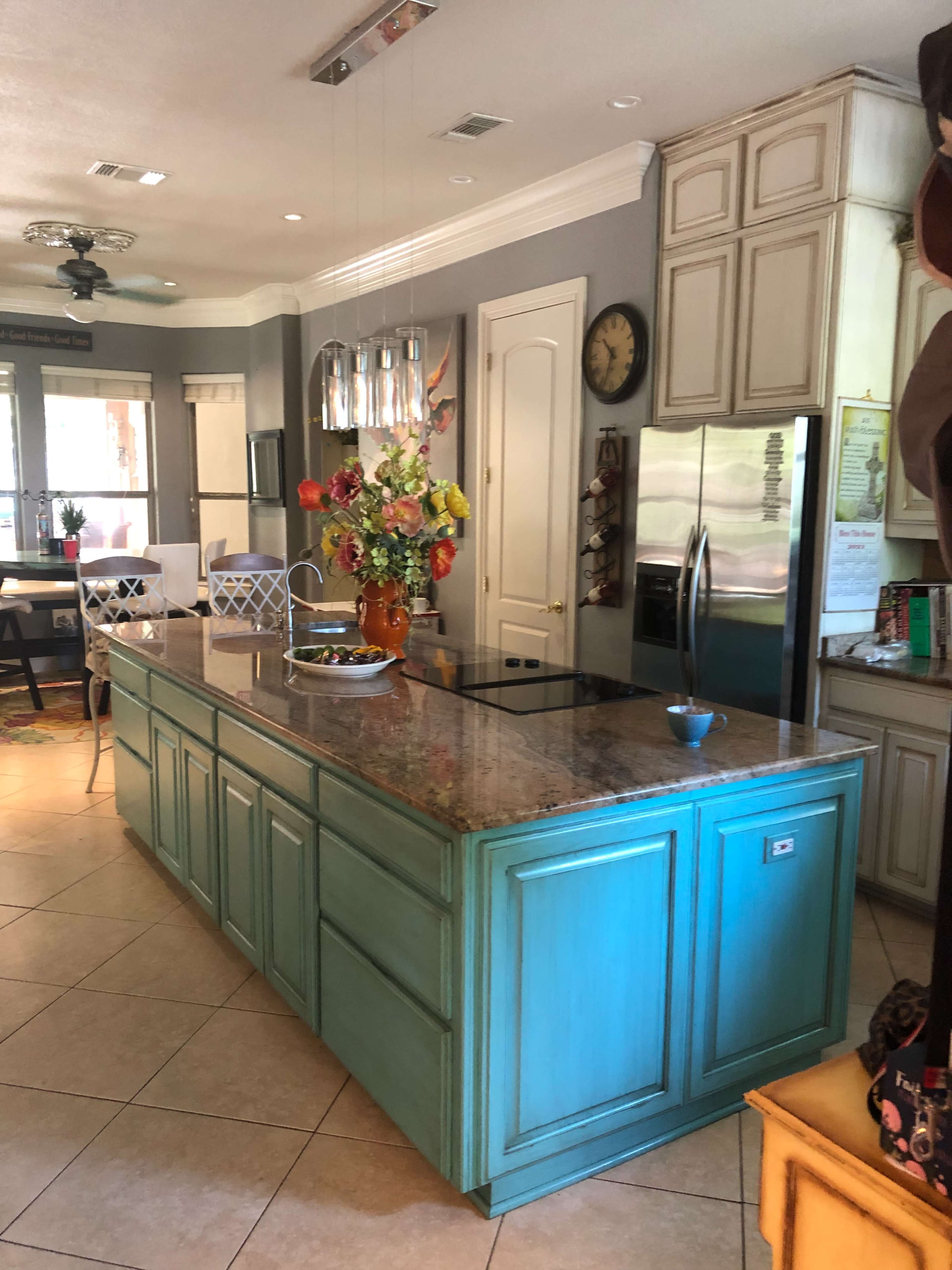 The kitchen features an island with a bright turquoise finish, a granite countertop, and various appliances, surrounded by light-colored cabinetry and a dining area in the background.
